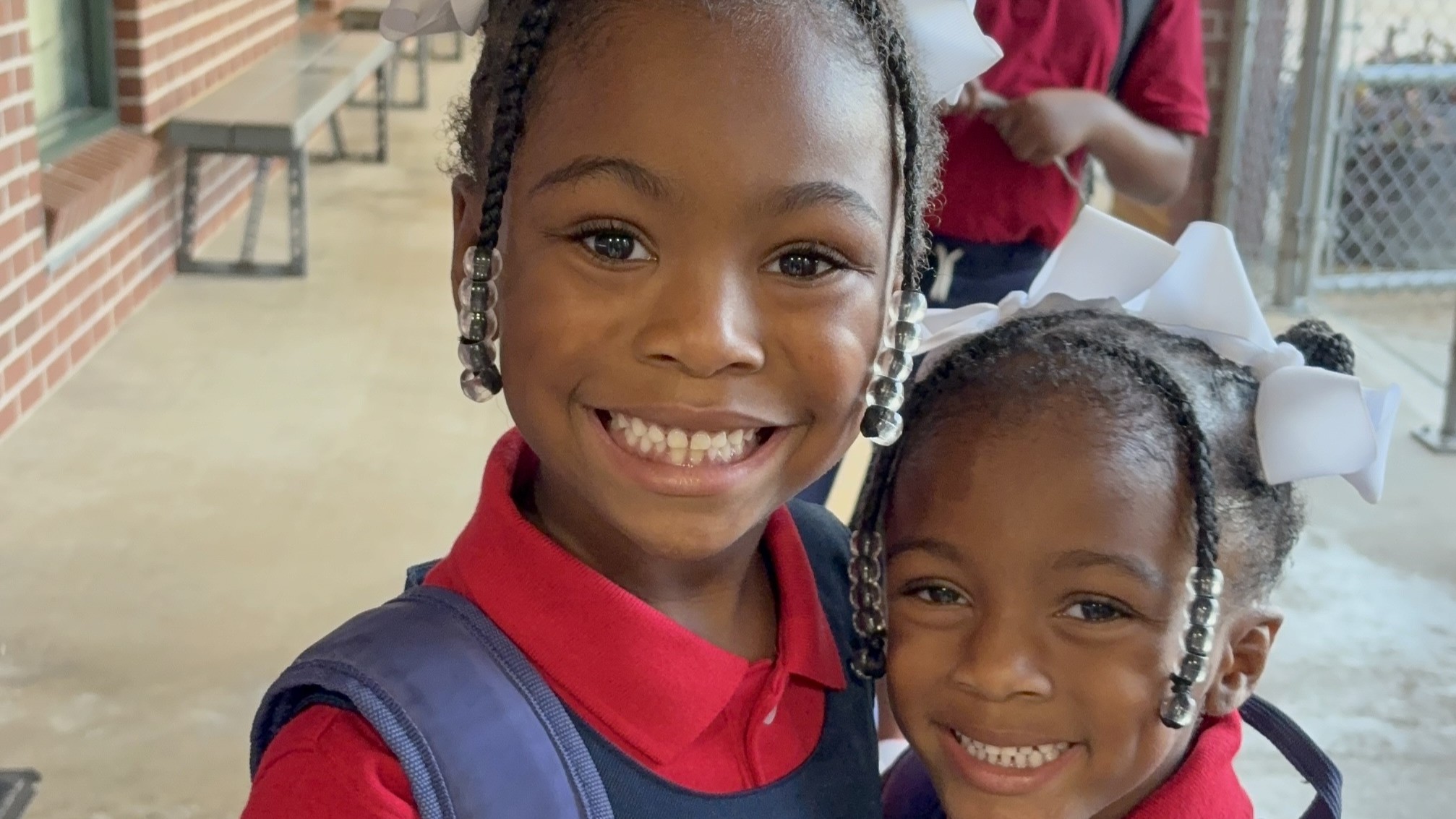 Two smiling young girls with braided hair and white bows are in the foreground, while a brick building and outdoor benches are visible in the background.