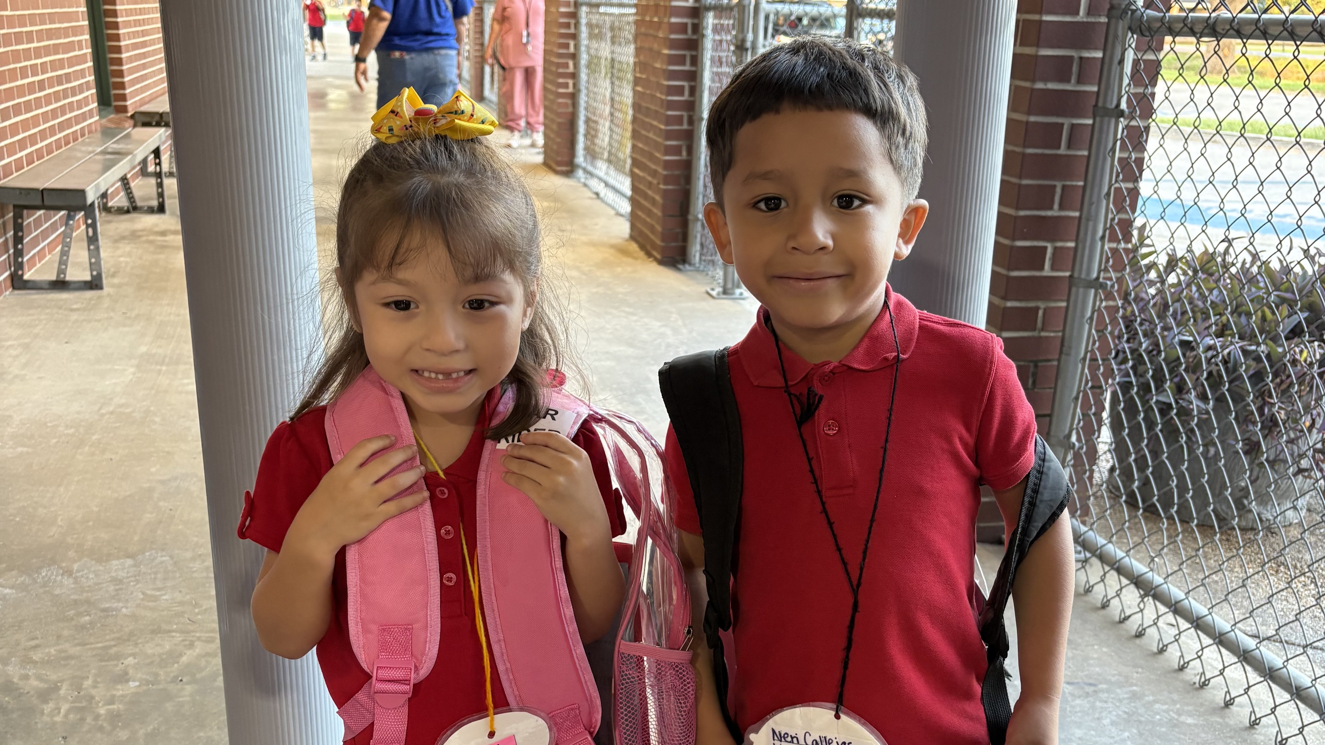 Two young children in red shirts and backpacks stand in a school hallway with a brick wall and outdoor greenery visible in the background.
