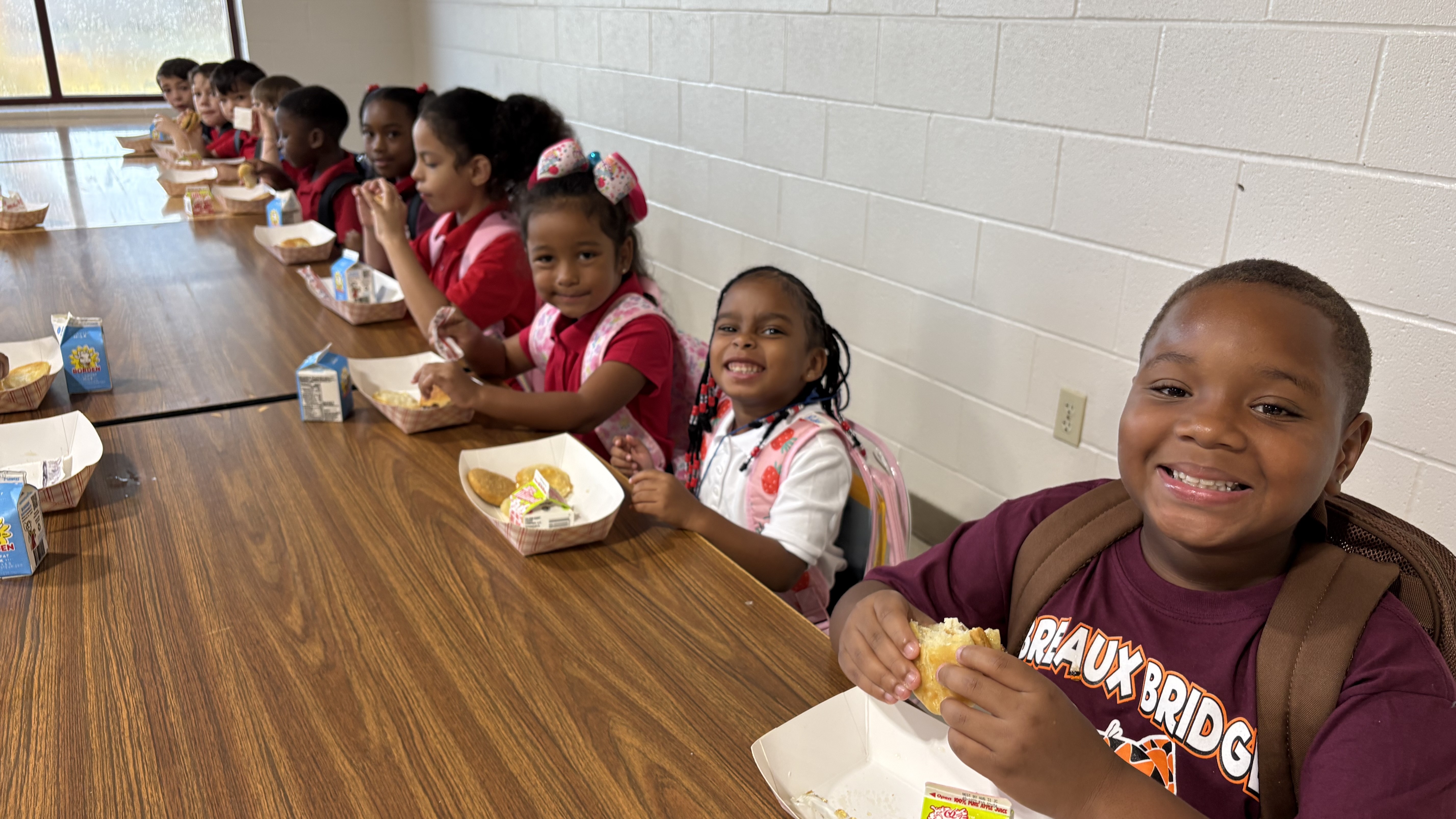 A group of smiling children in school uniforms sit at a long table eating lunch, with a plain wall in the background.