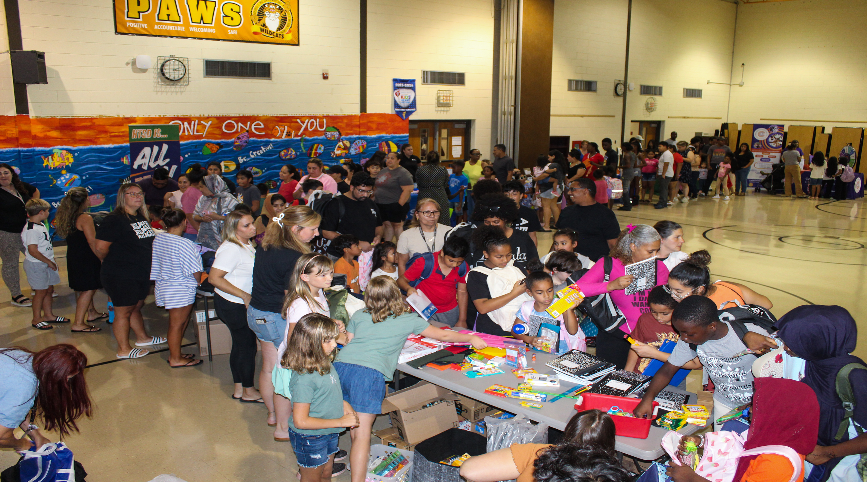 In the foreground, children and adults gather around tables laden with supplies, while the background reveals a gymnasium with a mural and banners.