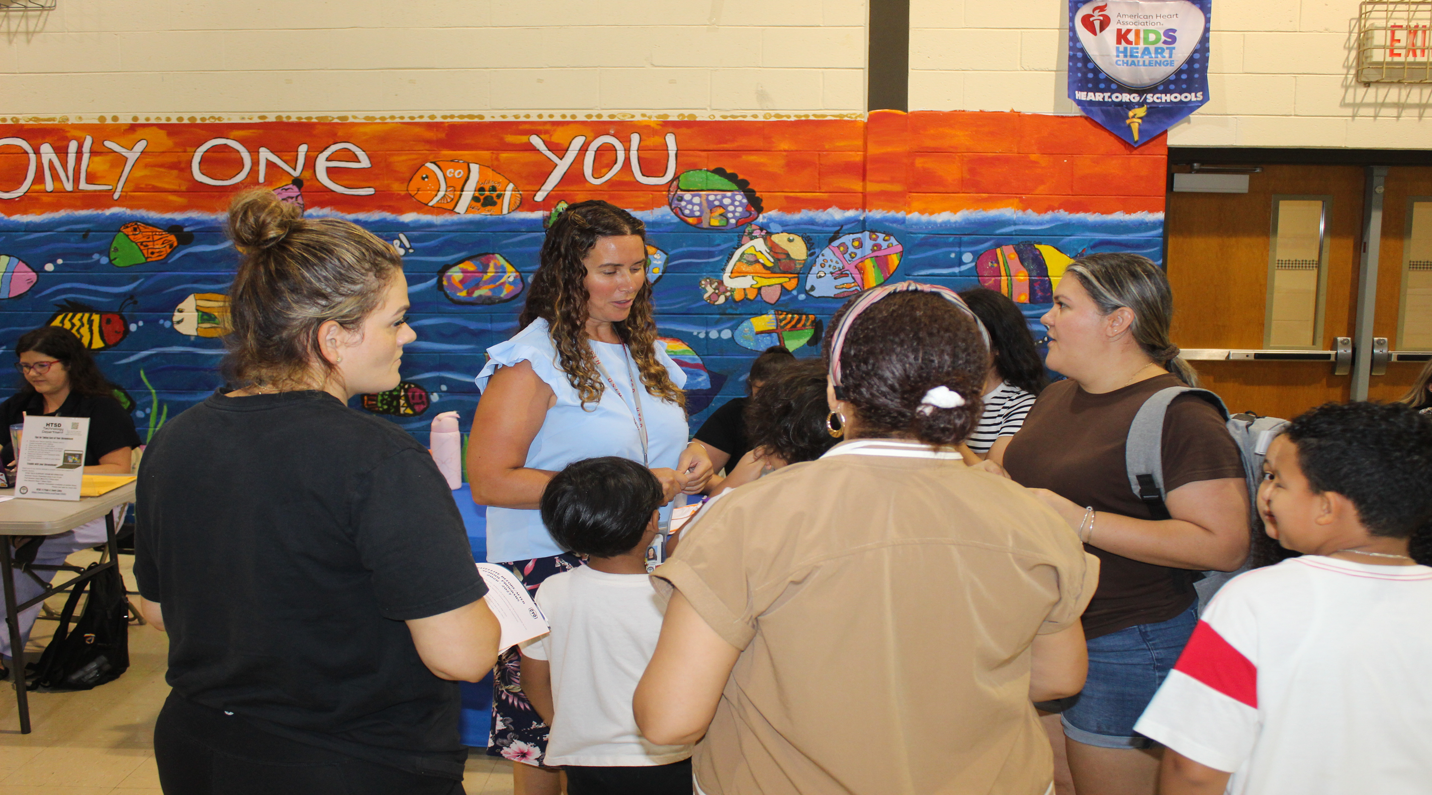 A group of adults and children gather in front of a colorful mural that reads "ONLY ONE YOU" in a school gymnasium.