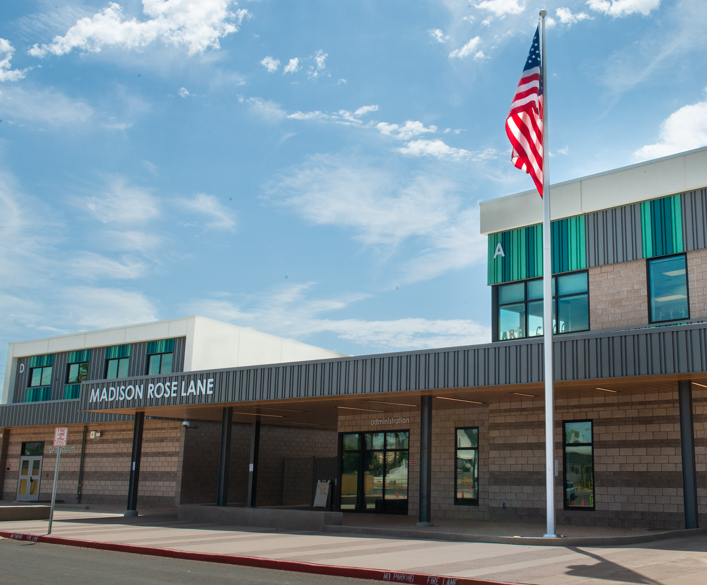 A flagpole with an American flag stands in front of a modern school building with a covered walkway and a bright blue sky overhead.