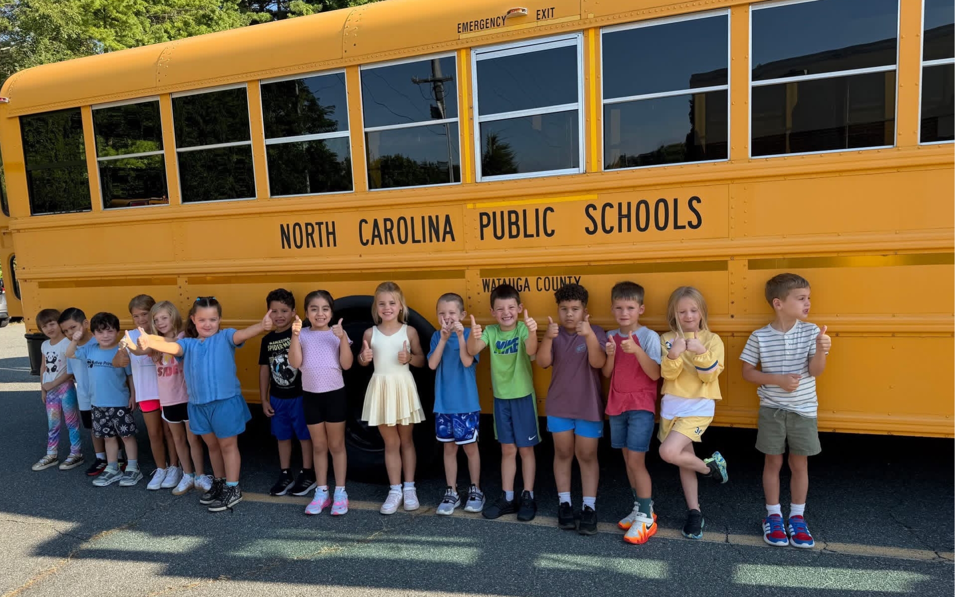 A group of elementary students smiling and giving a thumbs up motion in front of a yellow school bus.