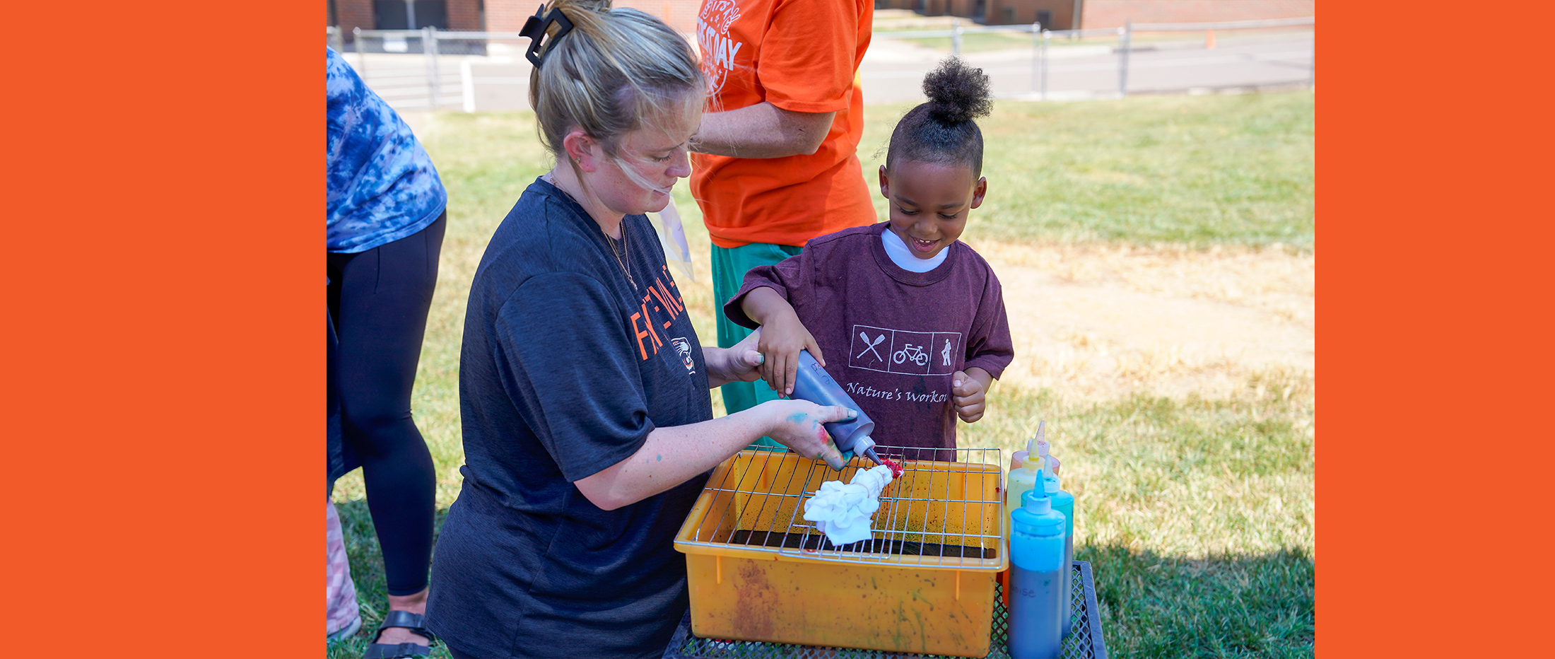 A woman and a young child are tie-dyeing a shirt outdoors with a grassy field and building in the background.