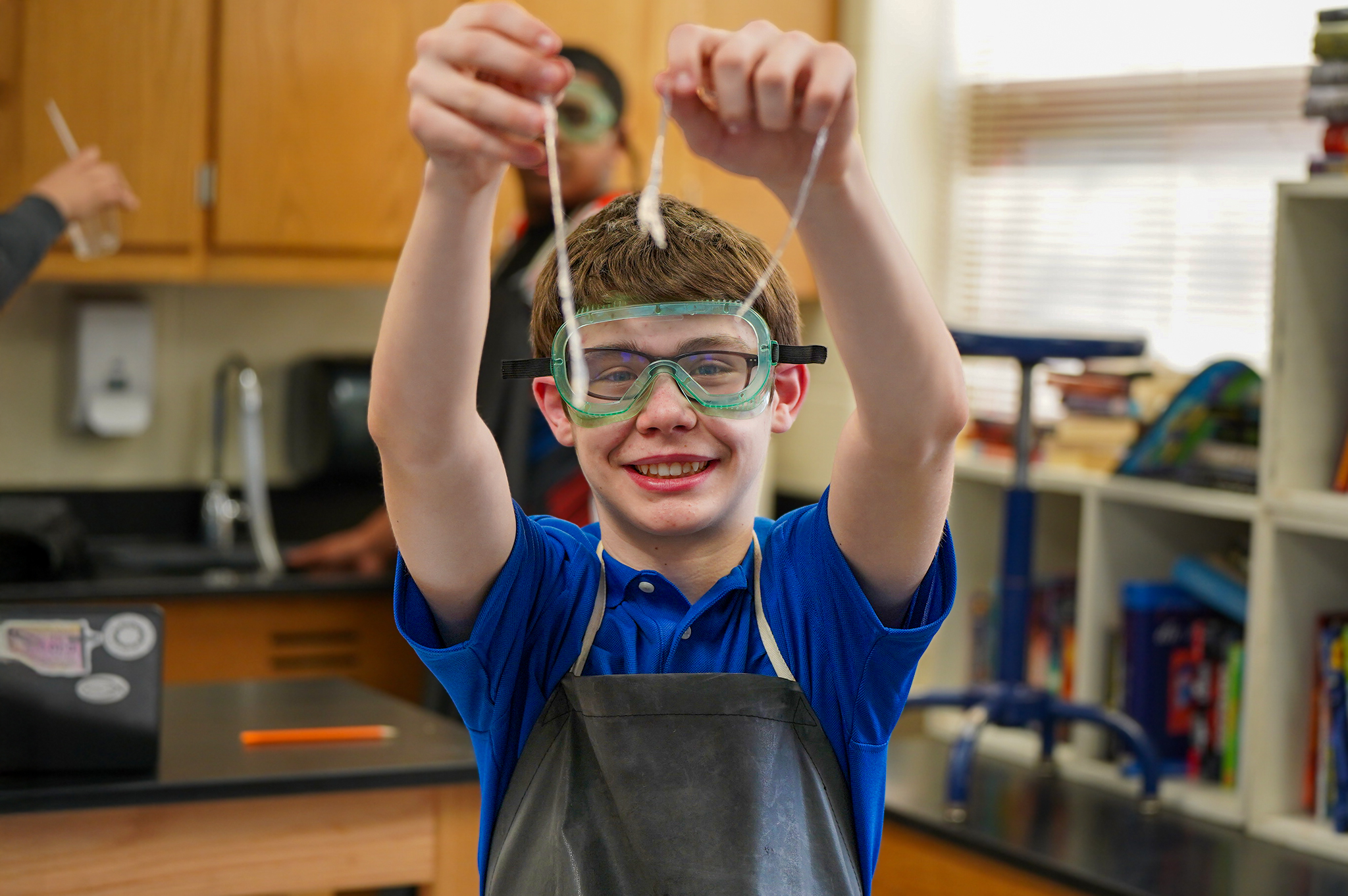A young boy in safety goggles and an apron holds up two cotton swabs in a classroom laboratory with cabinets and shelves in the background.