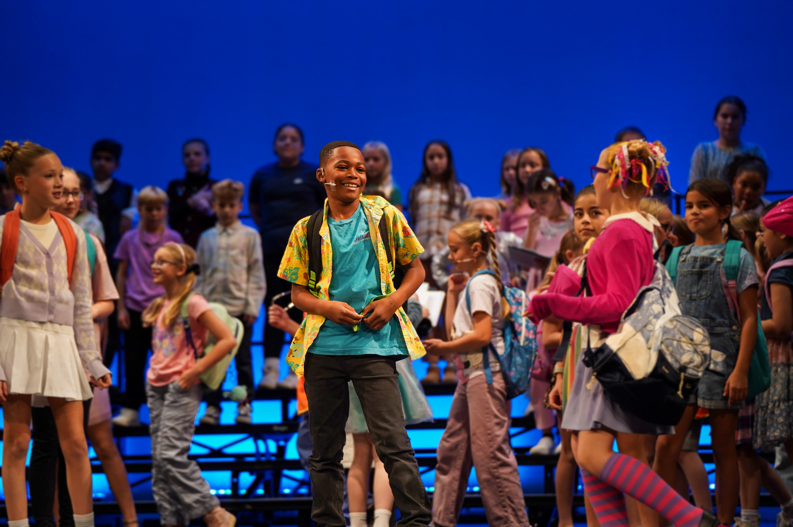 A young boy in a colorful shirt smiles in the foreground, while a diverse group of children stands on risers against a bright blue background.
