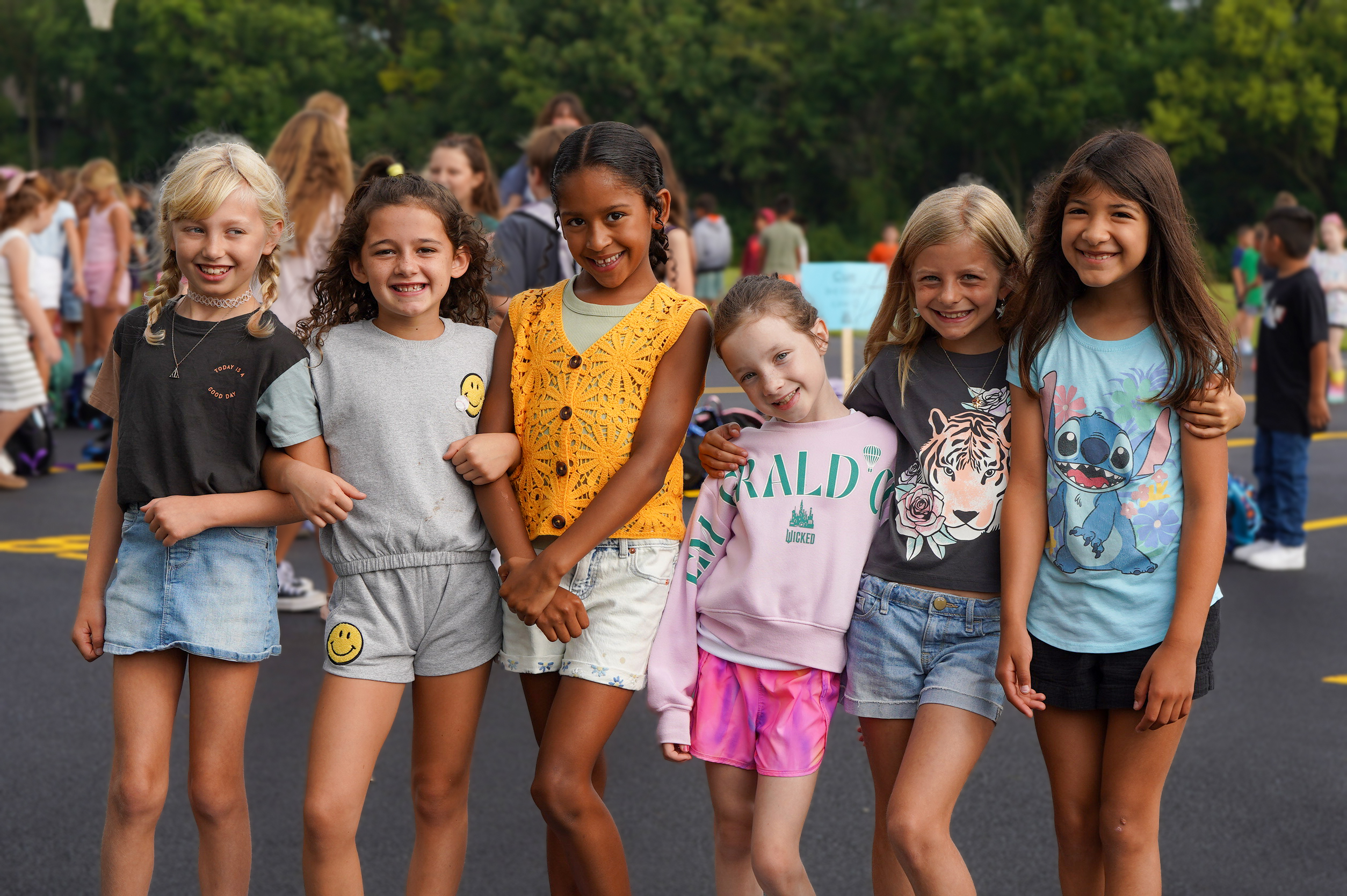 Six smiling young girls in casual summer clothes pose in the foreground with a blurred background of other children and trees.