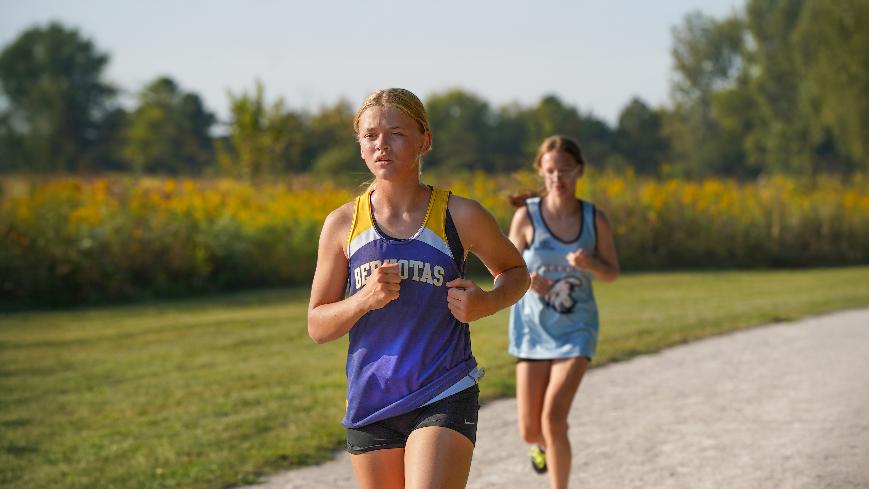 Two young women in running attire are jogging on a gravel path with a field of yellow flowers and trees in the background.