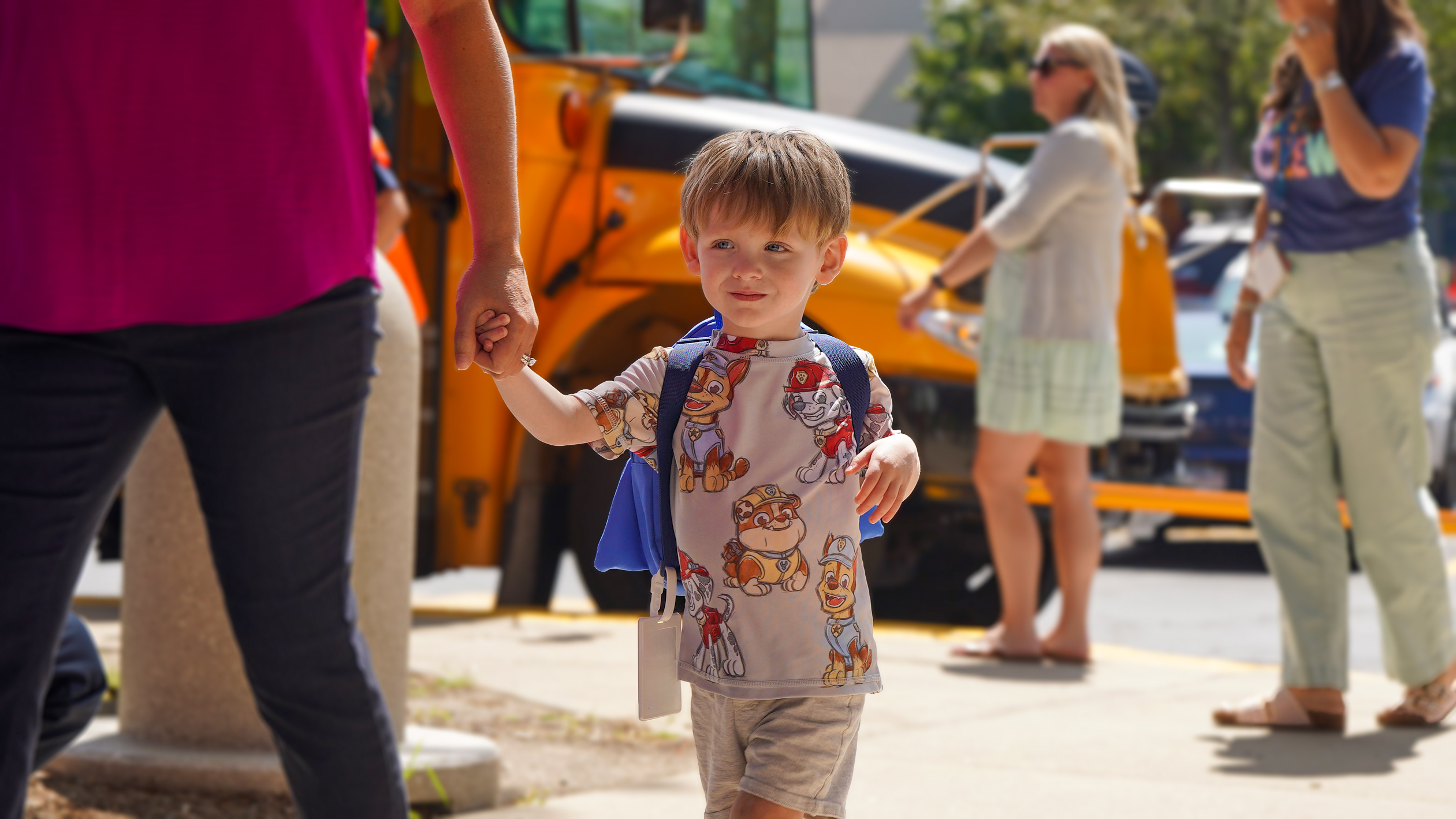 A young boy in a cartoon-patterned shirt and backpack holds an adult's hand as they walk past a yellow school bus and other people in the background.