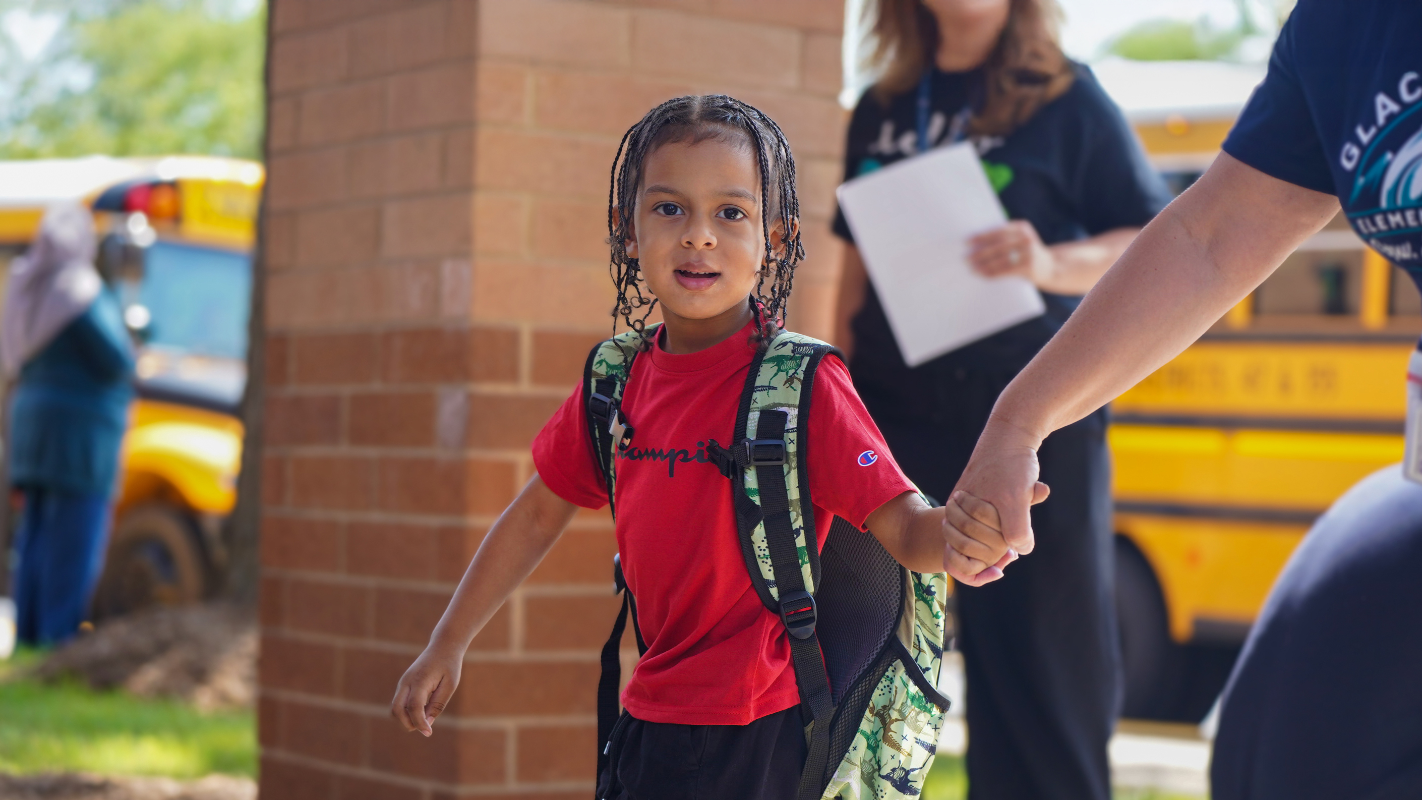 A young boy with braids and a red shirt, holding hands with an adult, walks towards a brick building with yellow school buses in the blurred background.