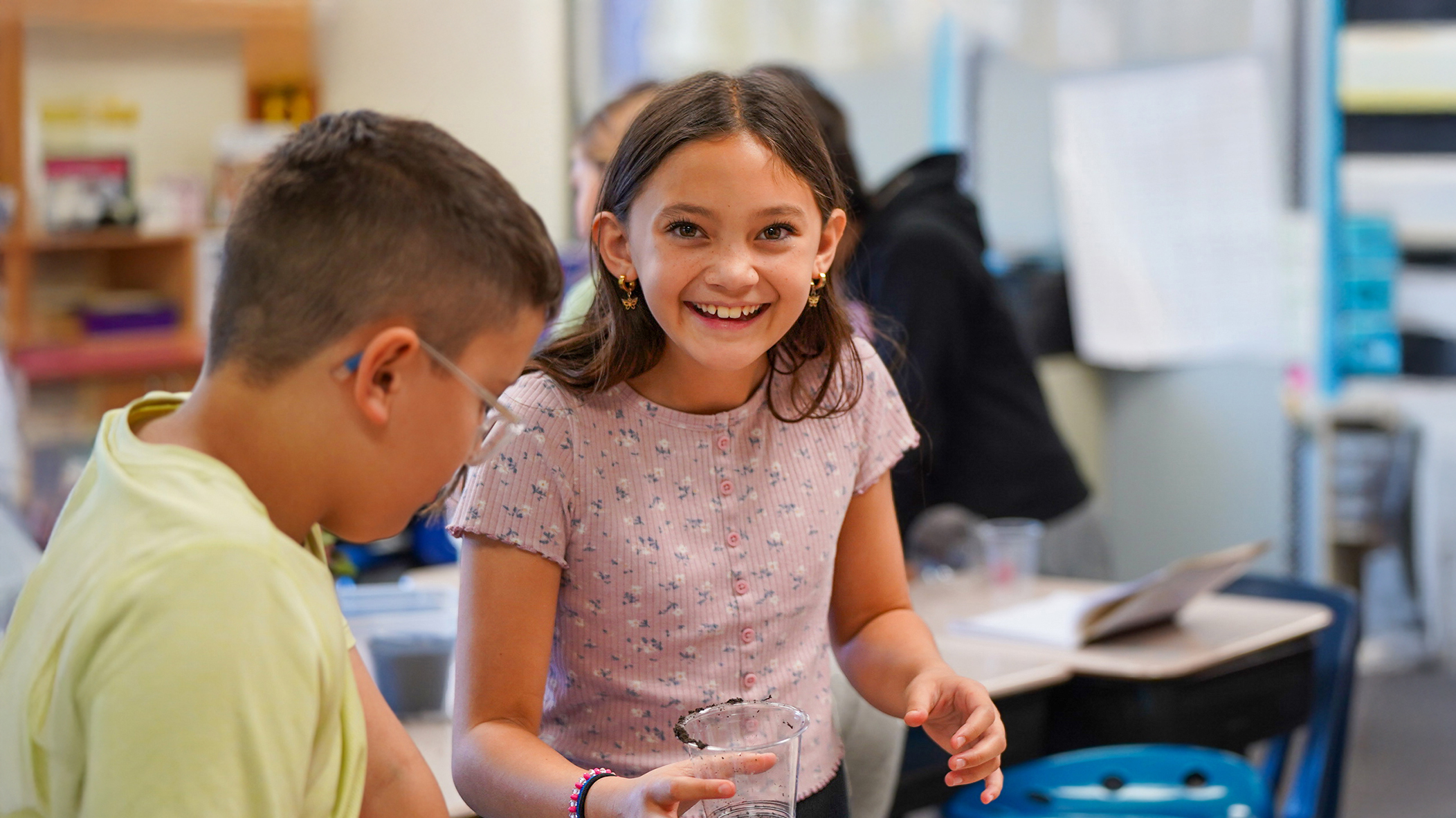 A smiling girl in the foreground is engaged in a science experiment with a classmate, while other students and classroom elements are blurred in the background.