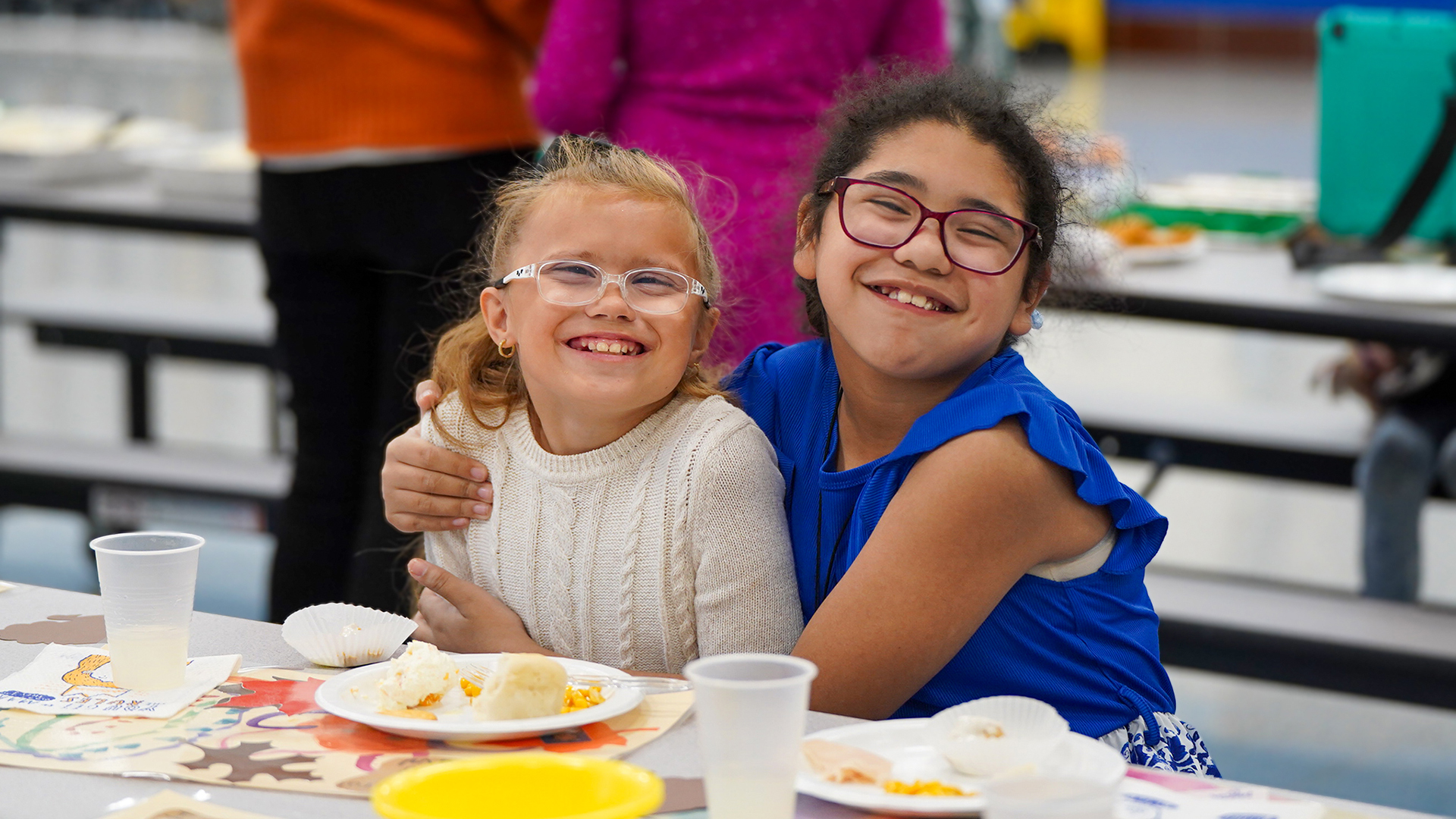 Two smiling girls with glasses embrace at a table with food and drinks, while blurred figures and a gymnasium are visible in the background.