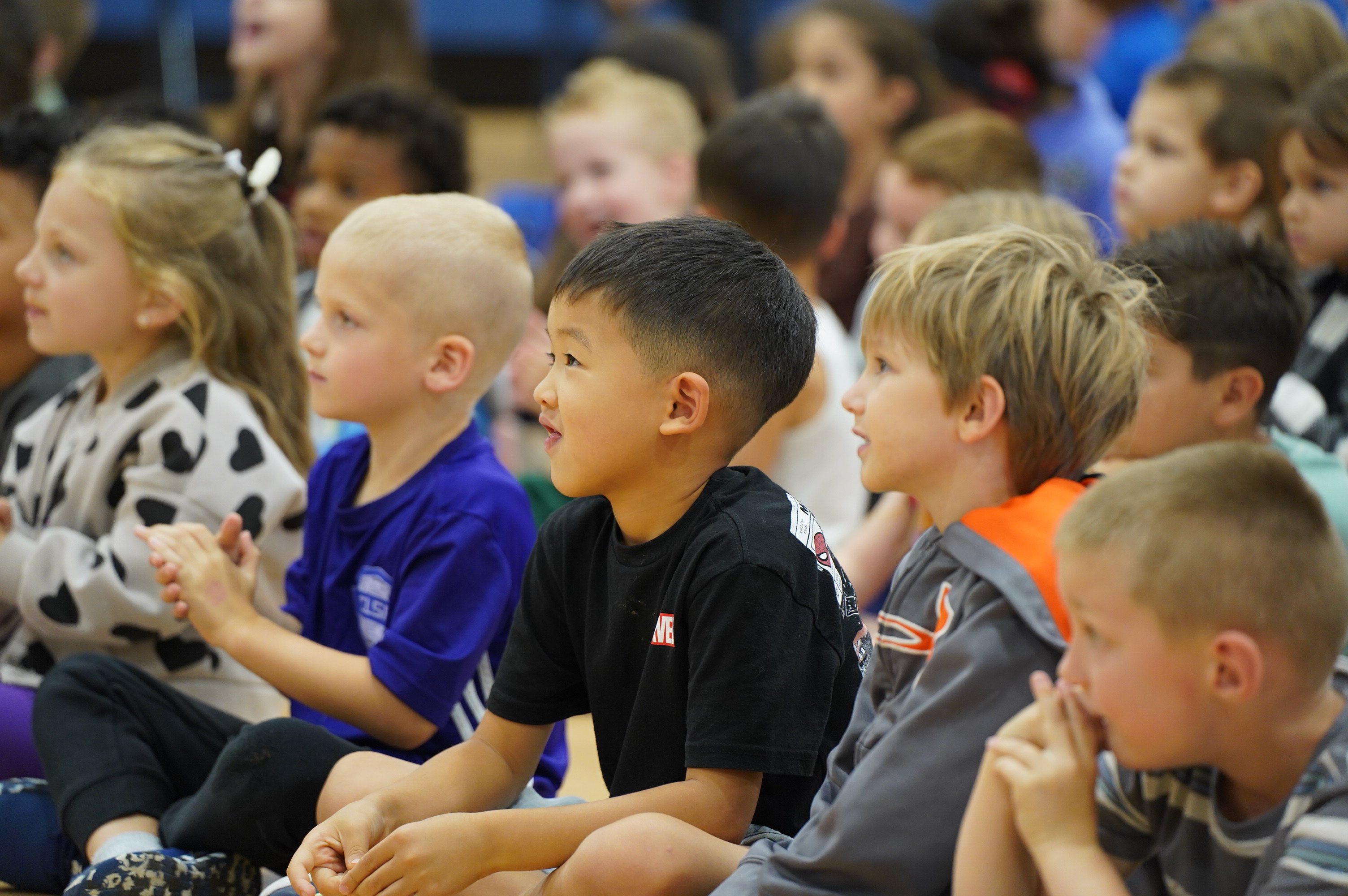 A group of young children, some clapping, sit attentively in the foreground, while others are visible in the blurred background.