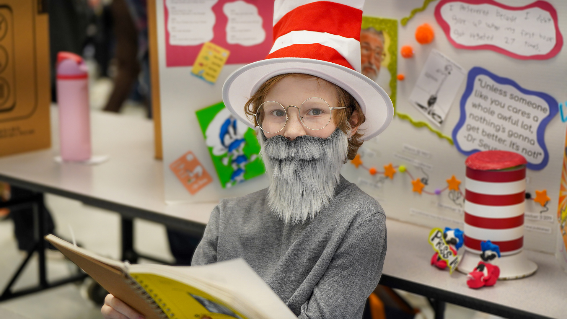 A young boy in a Cat in the Hat costume with a fake beard reads a book in the foreground, while colorful science fair projects and blurry figures fill the background.