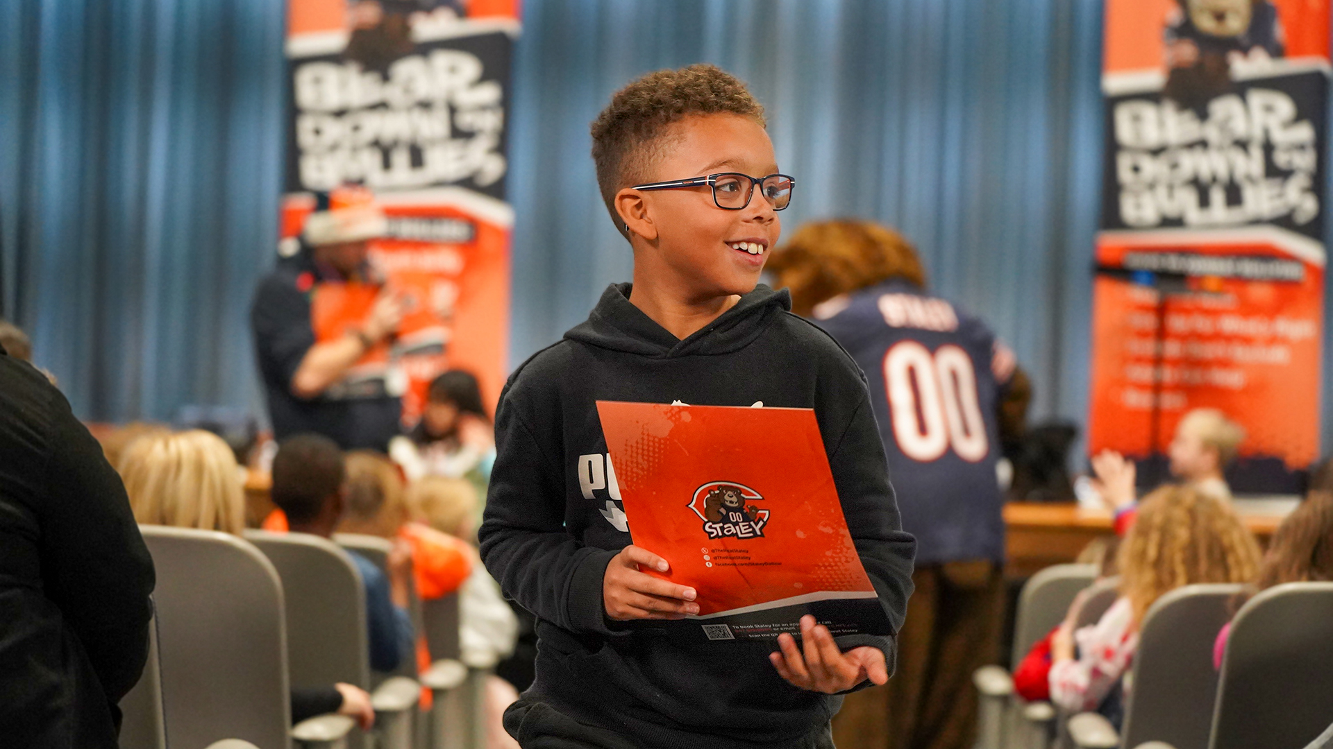 A young boy in glasses holds a folder as he walks past rows of seated children and adults, with banners in the background promoting "Bear Down for Bullying."