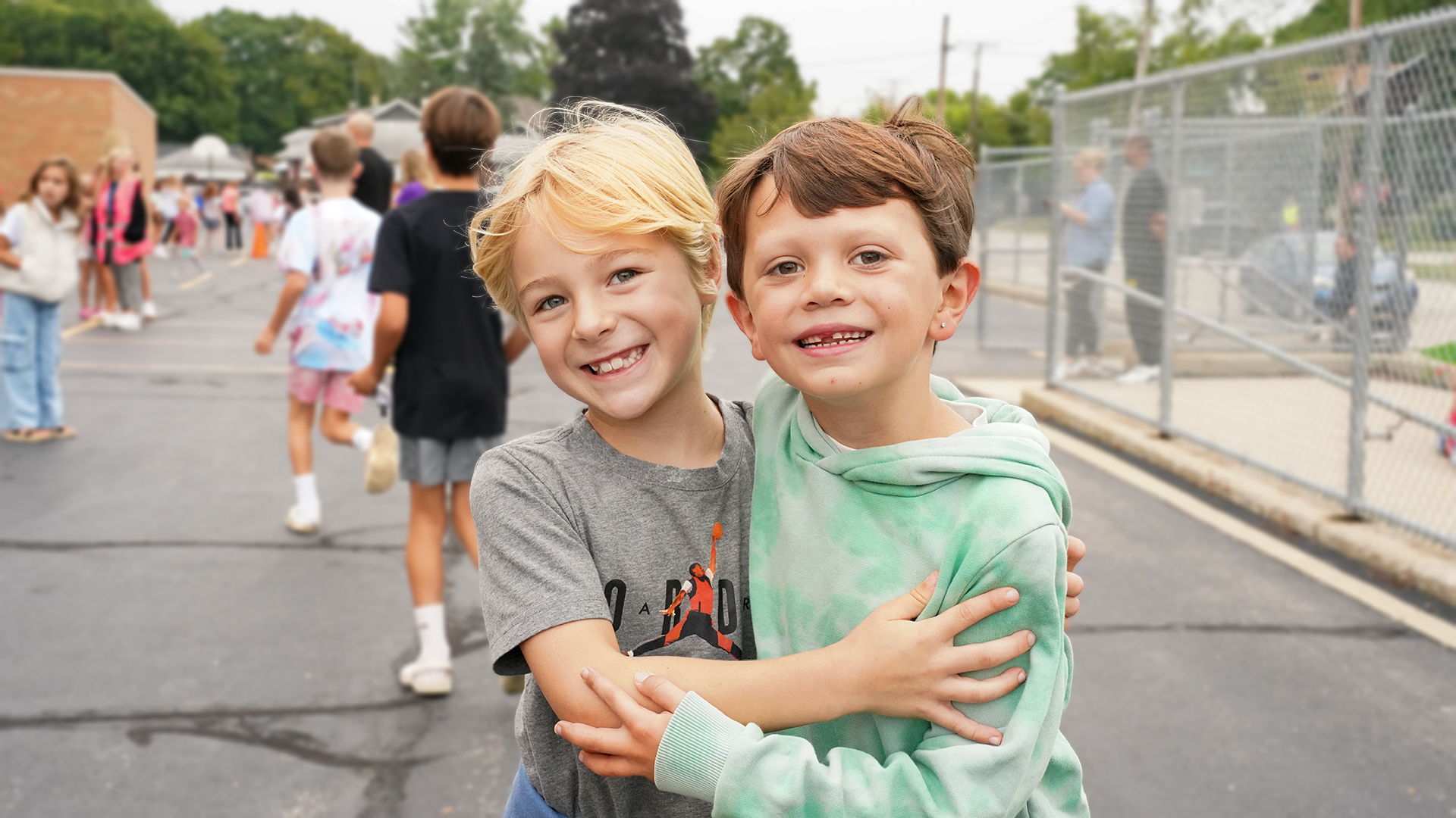Two smiling young boys embrace in the foreground, with a blurred background of other children and a school building.