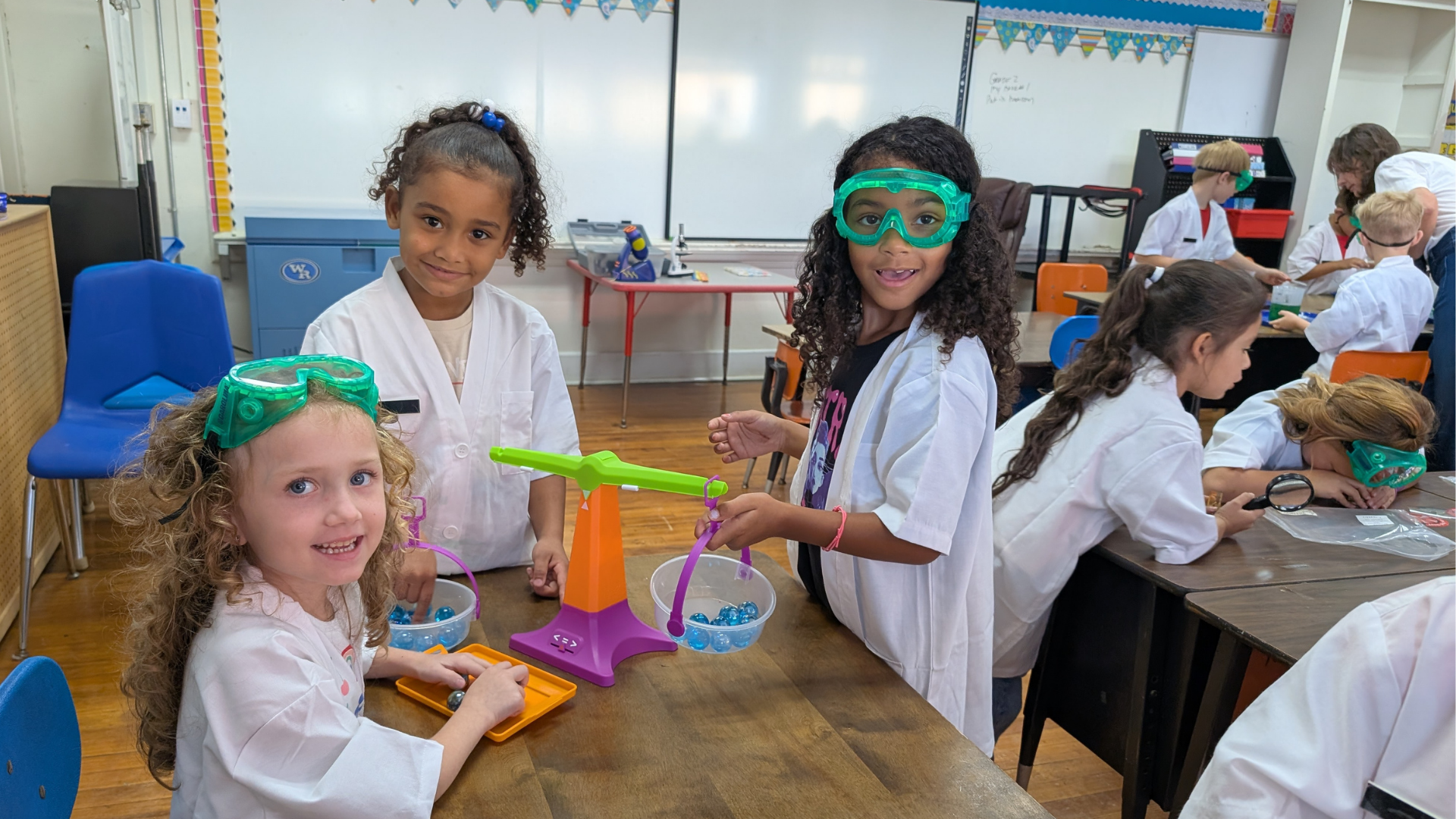 Three young girls in lab coats and goggles are engaged in a science experiment with a balance scale in the foreground, while other children in a classroom setting are visible in the background.
