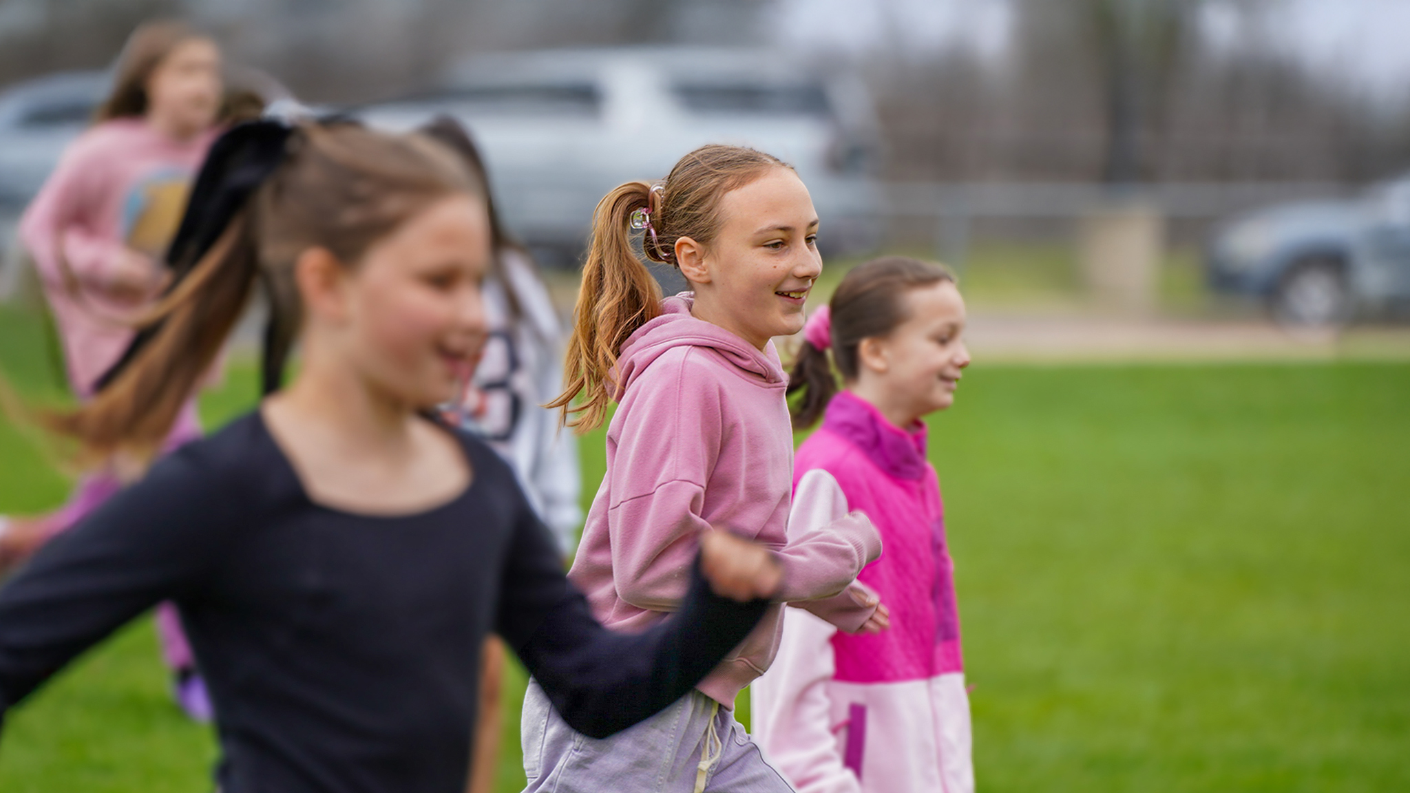 In the foreground, three young girls with ponytails run across a grassy field, while in the background, cars are parked under a cloudy sky.