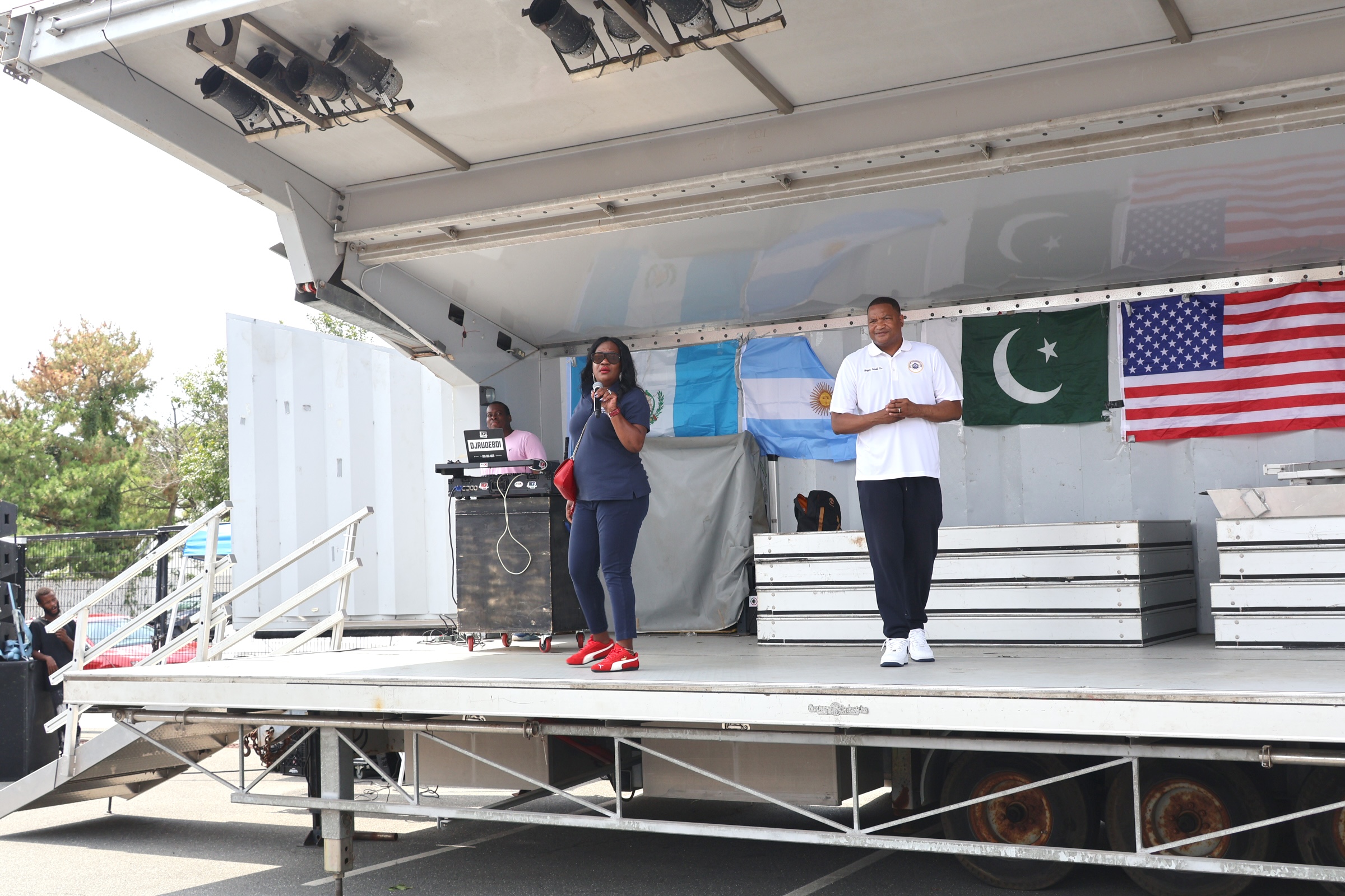 Two people stand on a stage with flags in the background, while a DJ booth is visible on the left side of the foreground.
