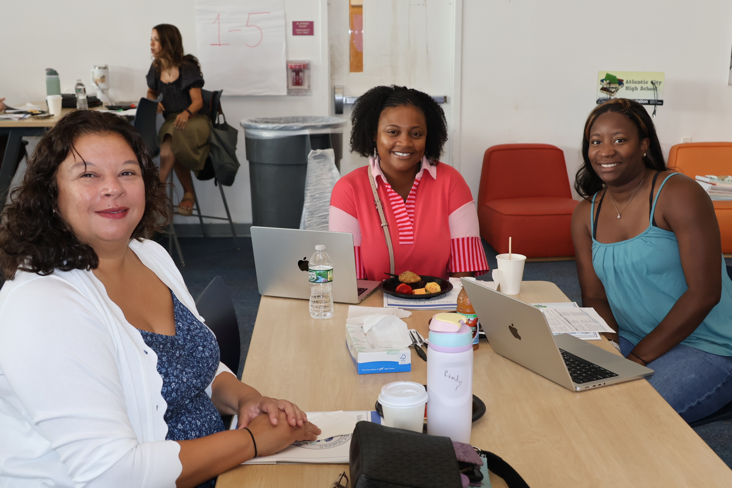 Three women sit at a table with laptops and snacks in the foreground, while a whiteboard and chairs are visible in the background.