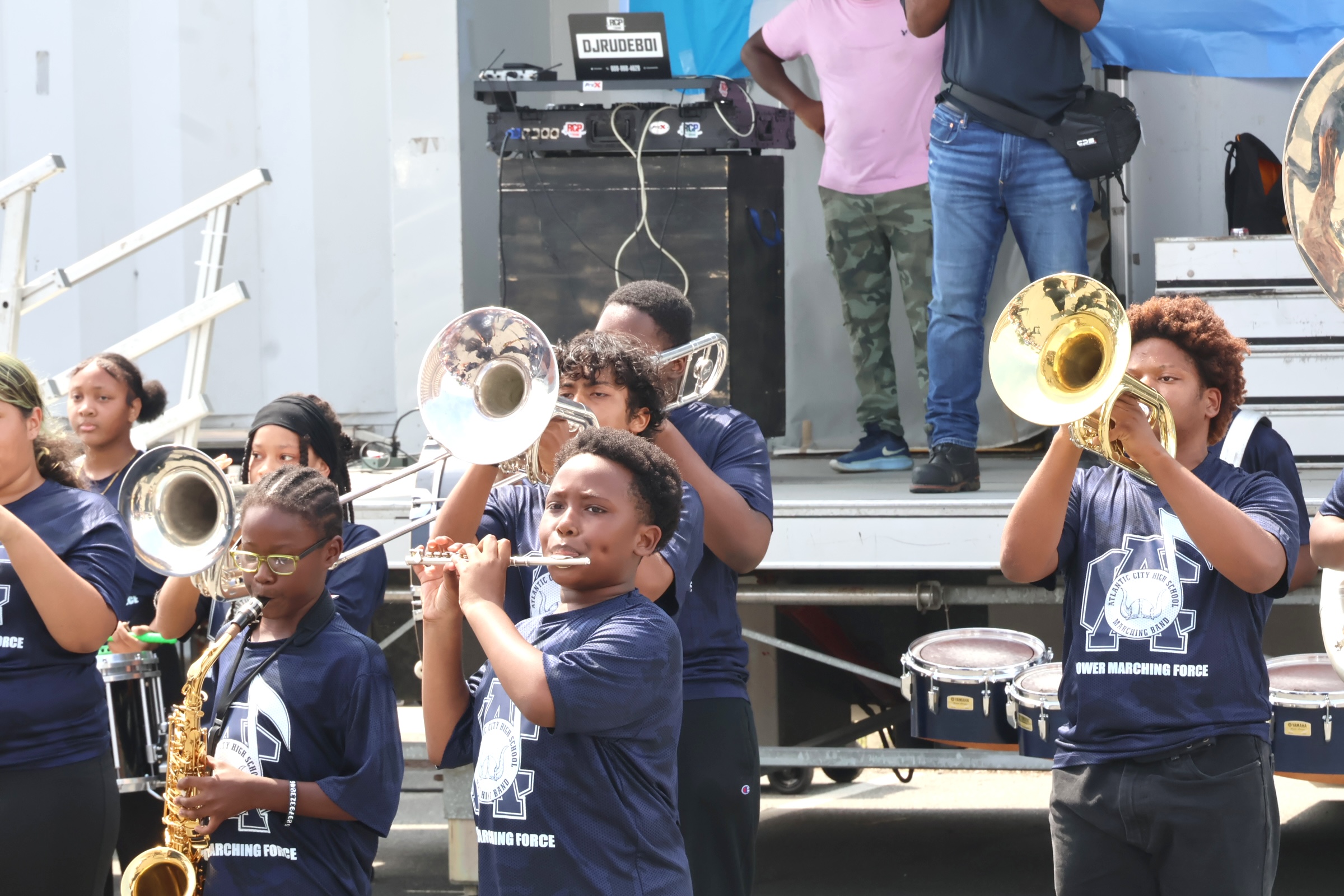 A group of young musicians in matching shirts play brass instruments and a flute in the foreground, with a DJ setup and spectators visible in the background.