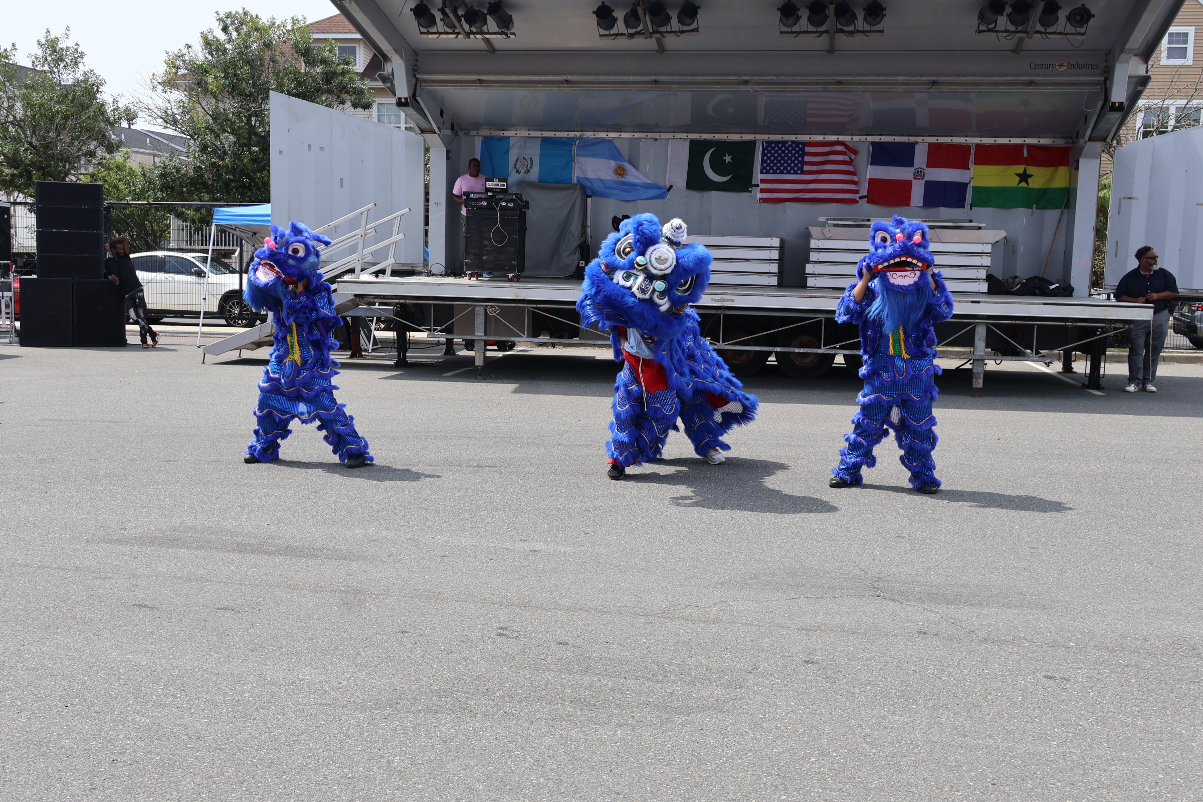 Three people in blue lion costumes dance on asphalt in front of a stage displaying various flags.