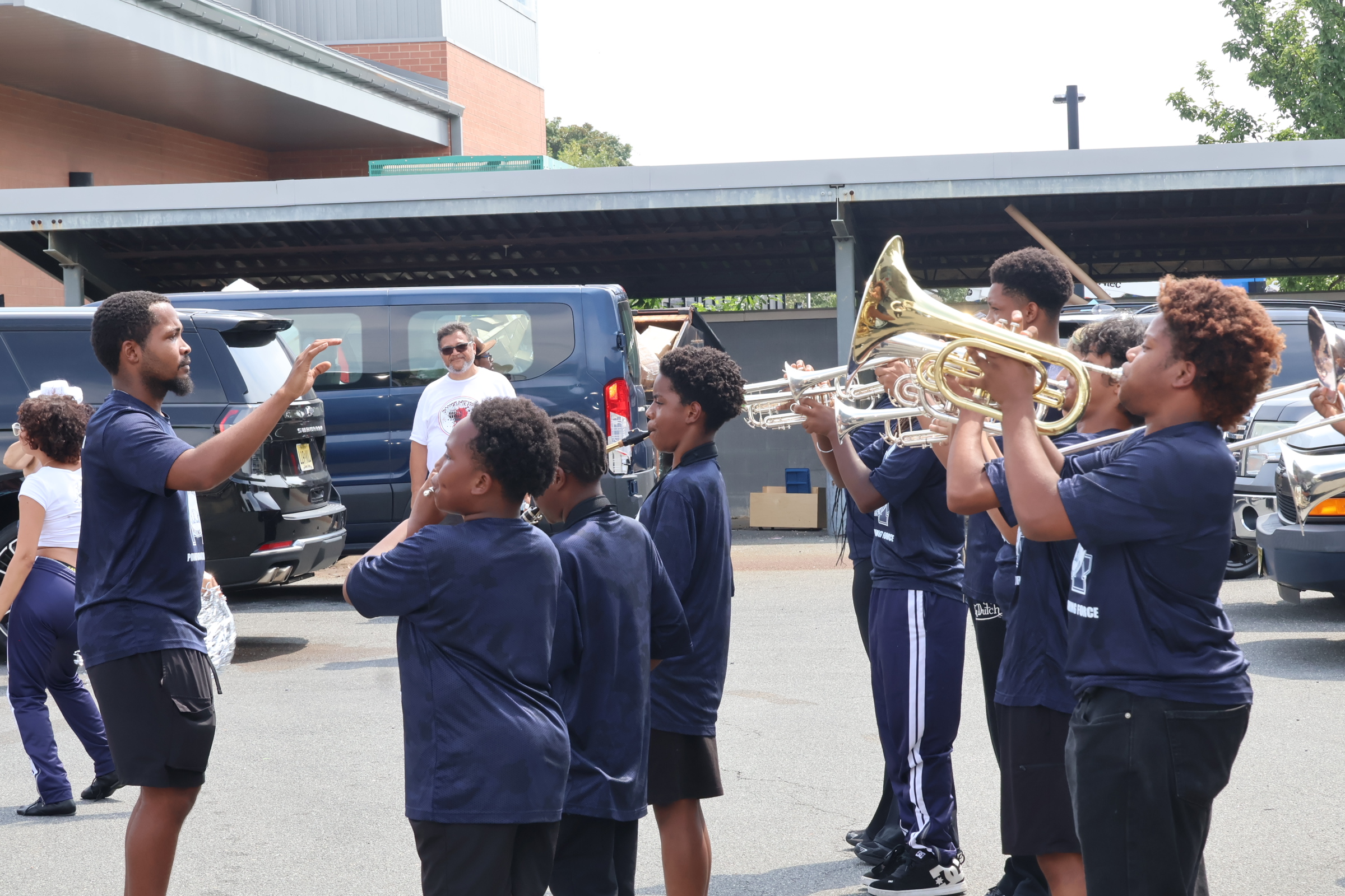 A band director leads a group of young musicians playing brass instruments in front of parked vans and a building.