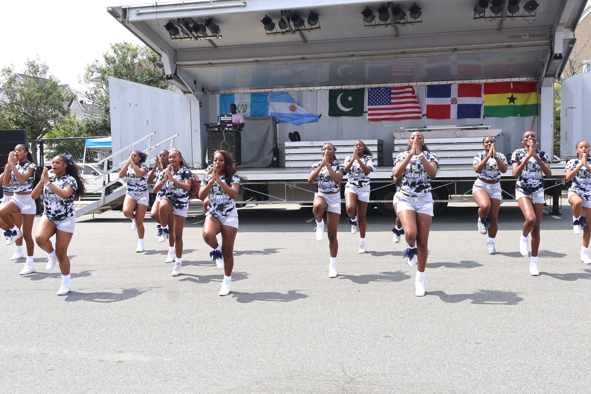 A group of young women in matching outfits perform a dance routine on a paved surface in front of a stage displaying several national flags.