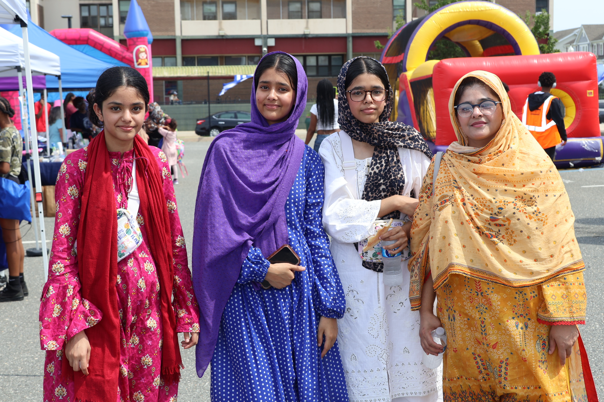 Four women in traditional attire stand in the foreground with inflatable attractions and tents in the background.