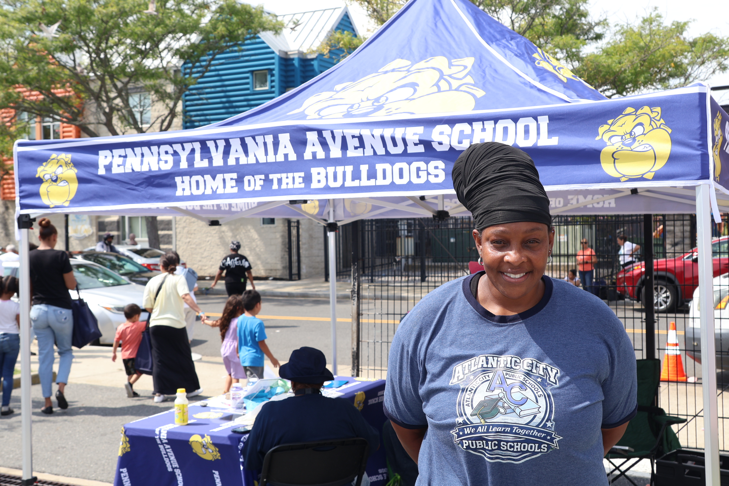 A woman stands in front of a tent for Pennsylvania Avenue School, with people and cars in the background.