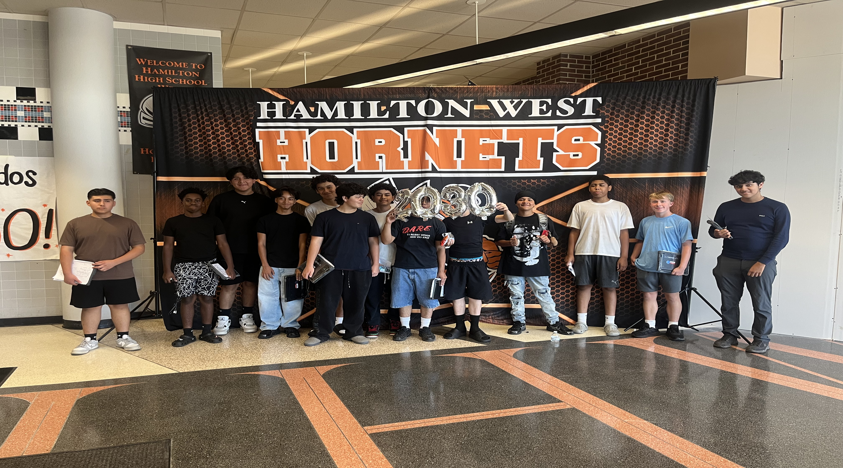 A group of young men stand in front of a "Hamilton-West Hornets" banner in a hallway, with a welcome sign visible in the background.