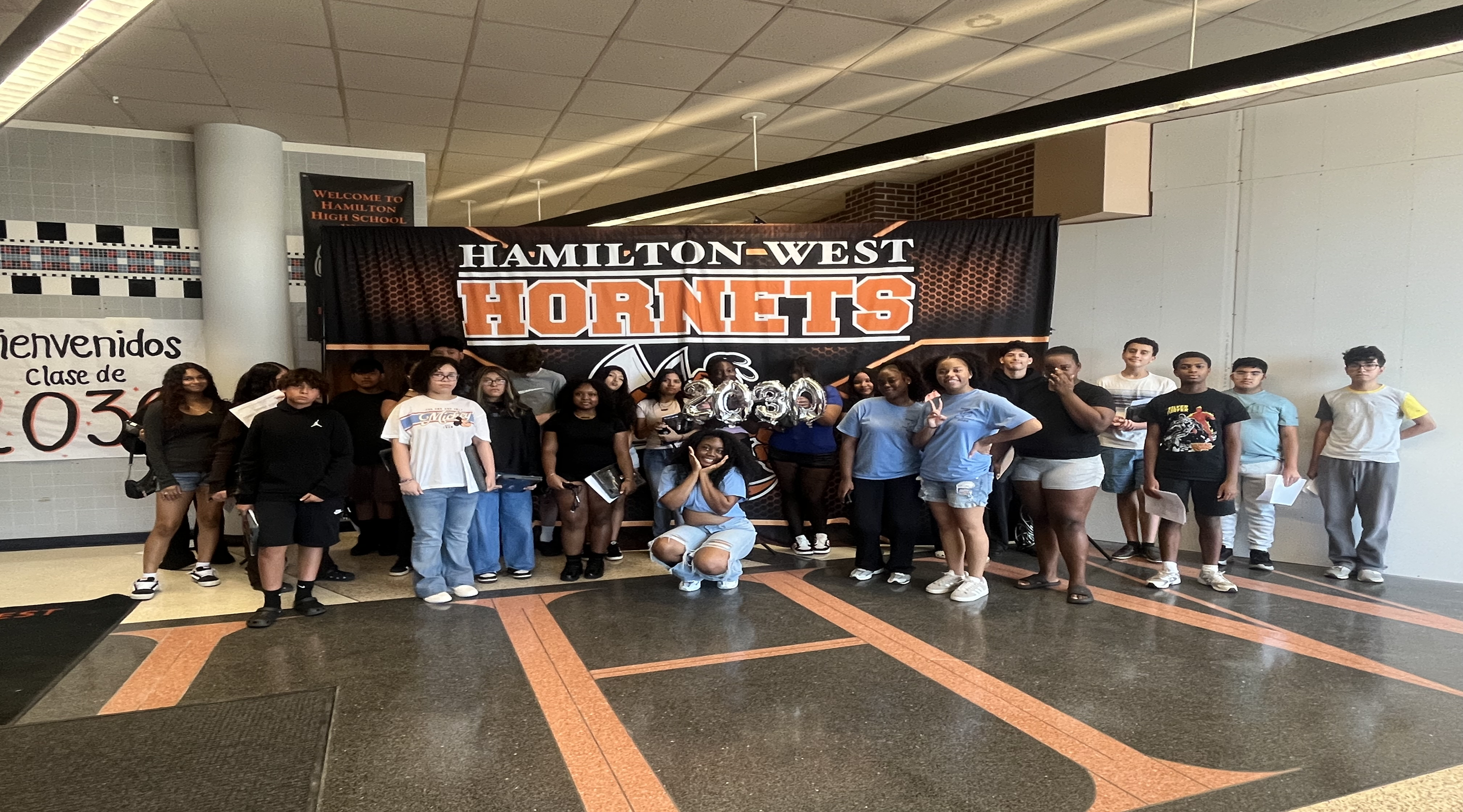 A group of students stands in a hallway in front of a "Hamilton-West Hornets" banner, with a large "H" design on the floor in the foreground.