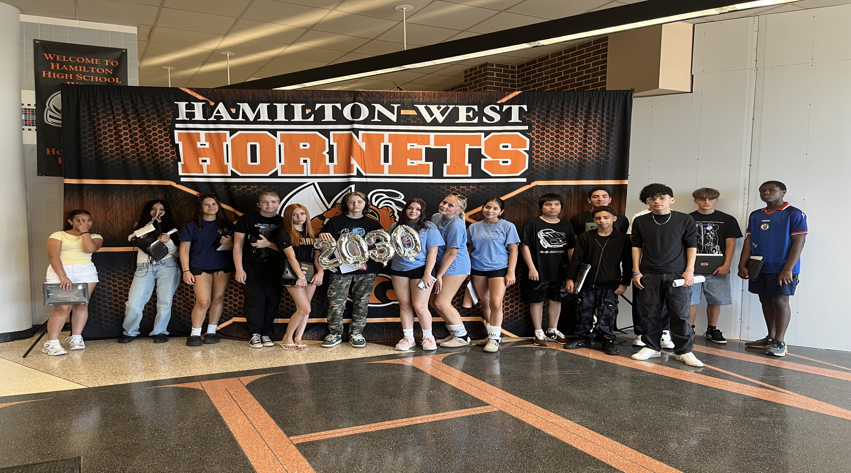 A group of teenagers poses for a photo in front of a "Hamilton-West Hornets" banner in a school hallway.