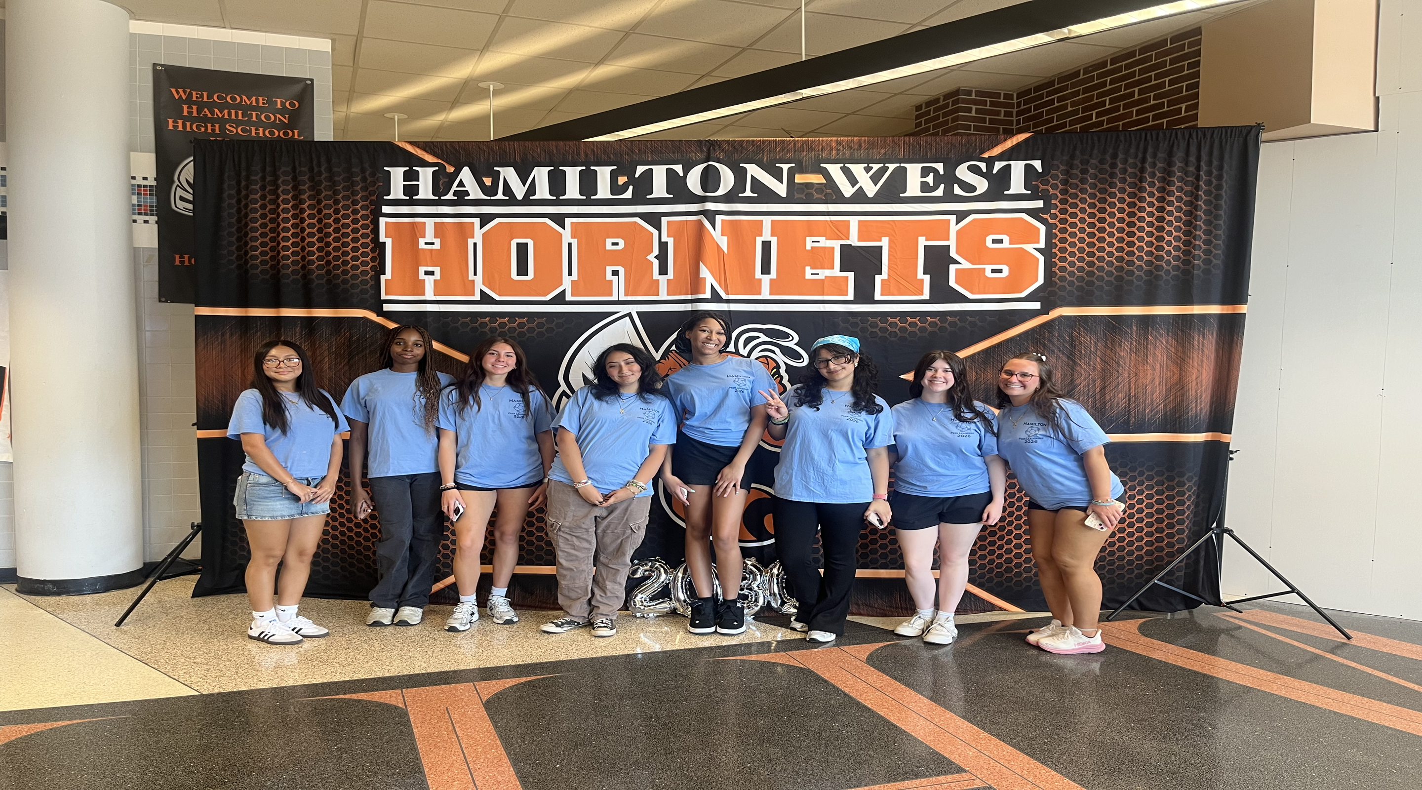 A group of young people in light blue shirts stand in front of a "Hamilton-West Hornets" banner, with a "Welcome to Hamilton High School" sign visible in the background.