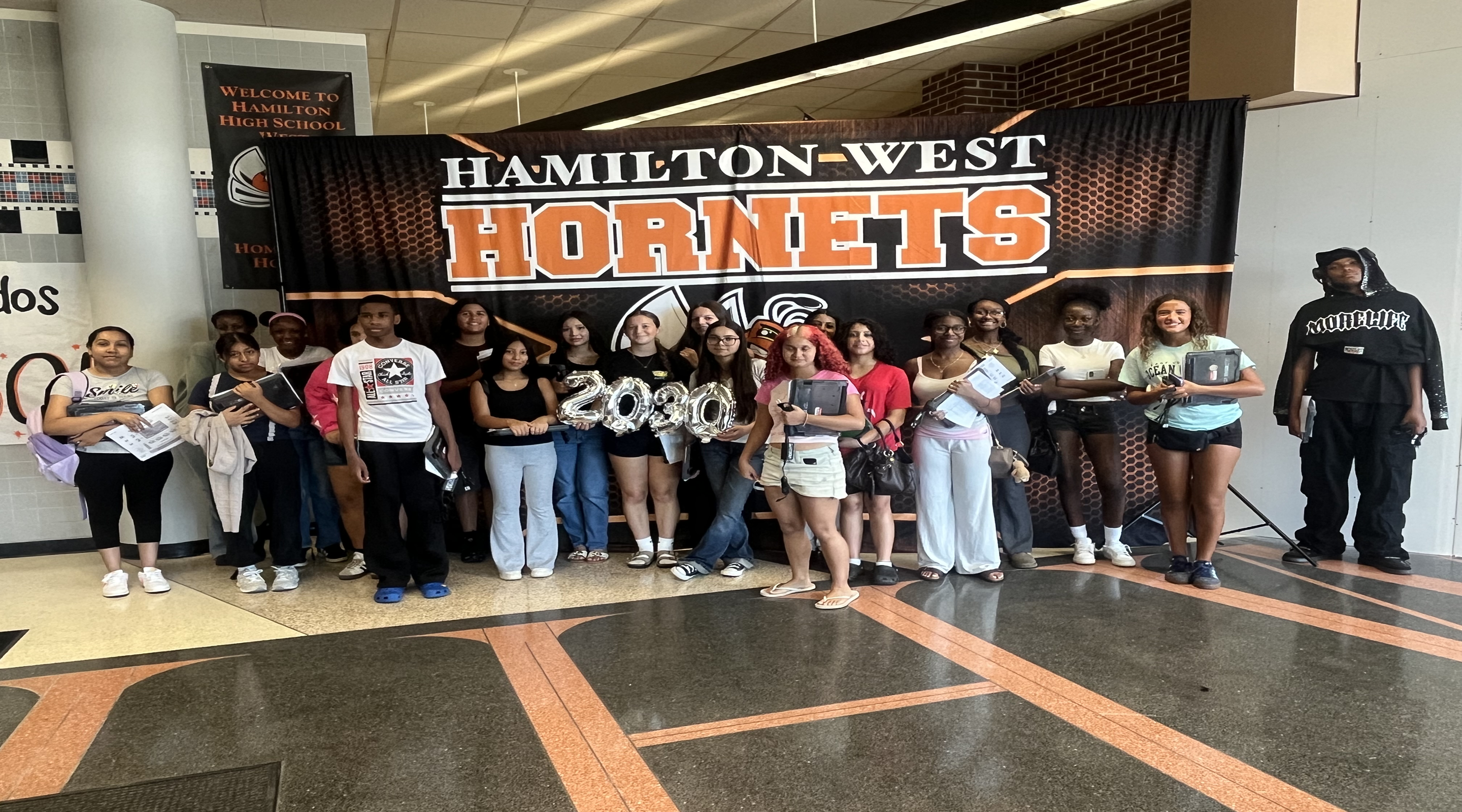 A group of students poses for a photo in front of a "Hamilton-West Hornets" banner, with a "Welcome to Hamilton High School" sign visible in the background.