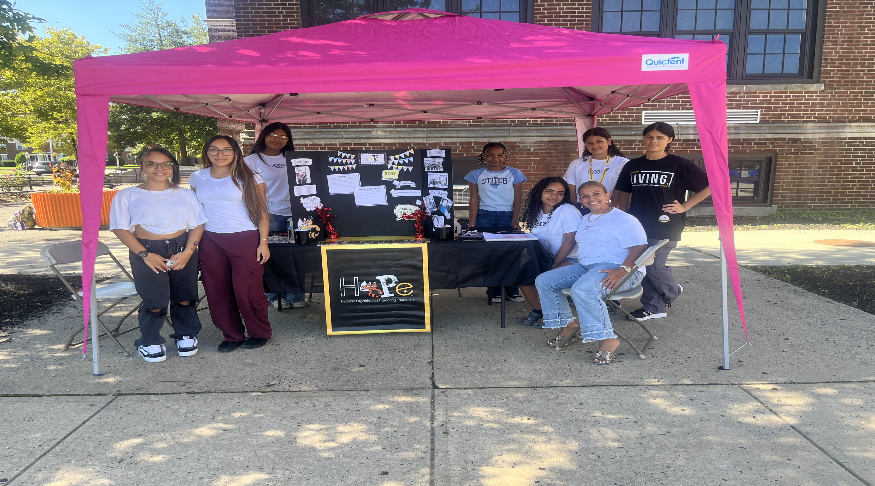 A group of young people stand under a bright pink tent in front of a brick building, with a table displaying information and a sign that says "HOPE."
