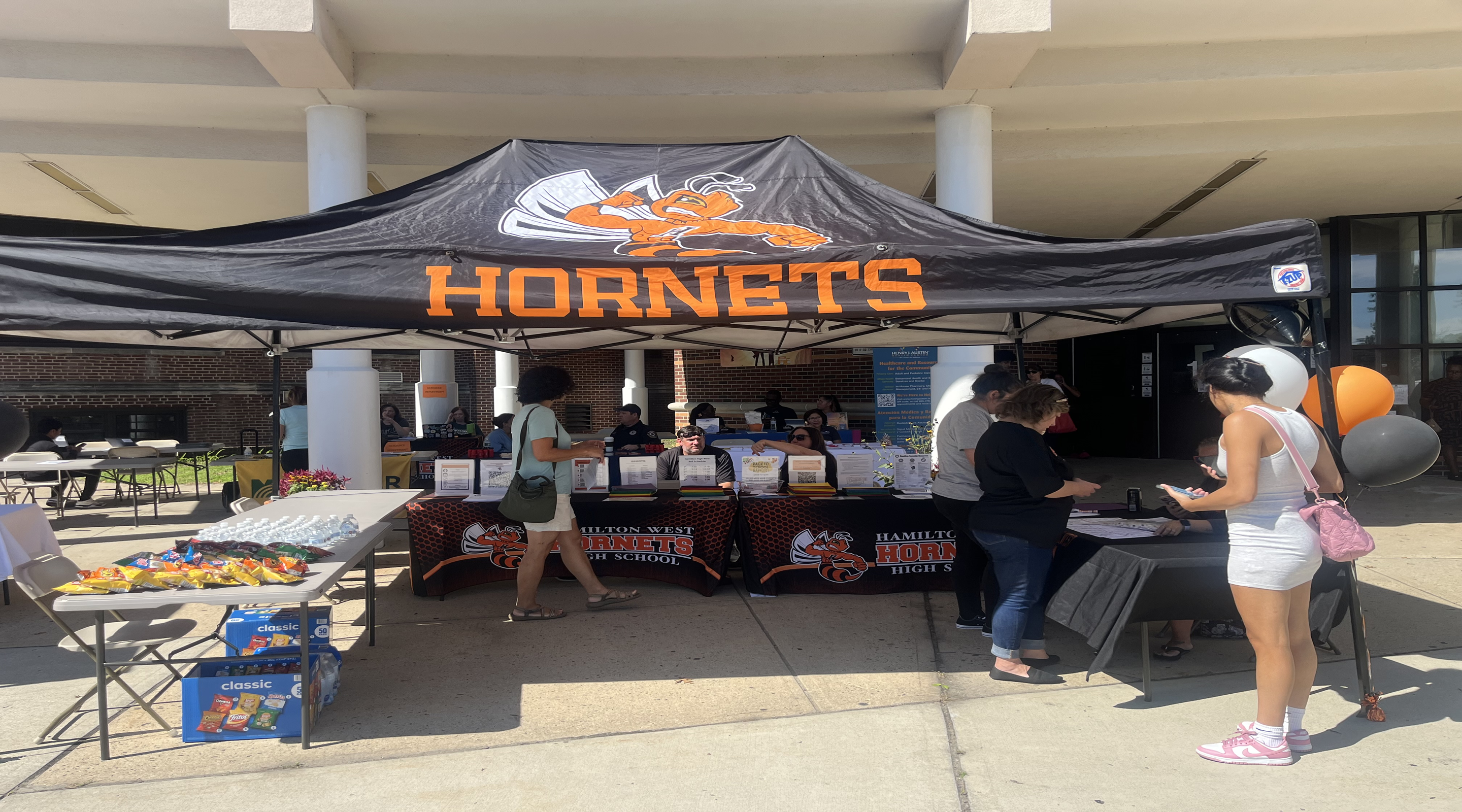 People gather under a "Hornets" tent with tables of snacks and information, while a building with columns stands in the background.