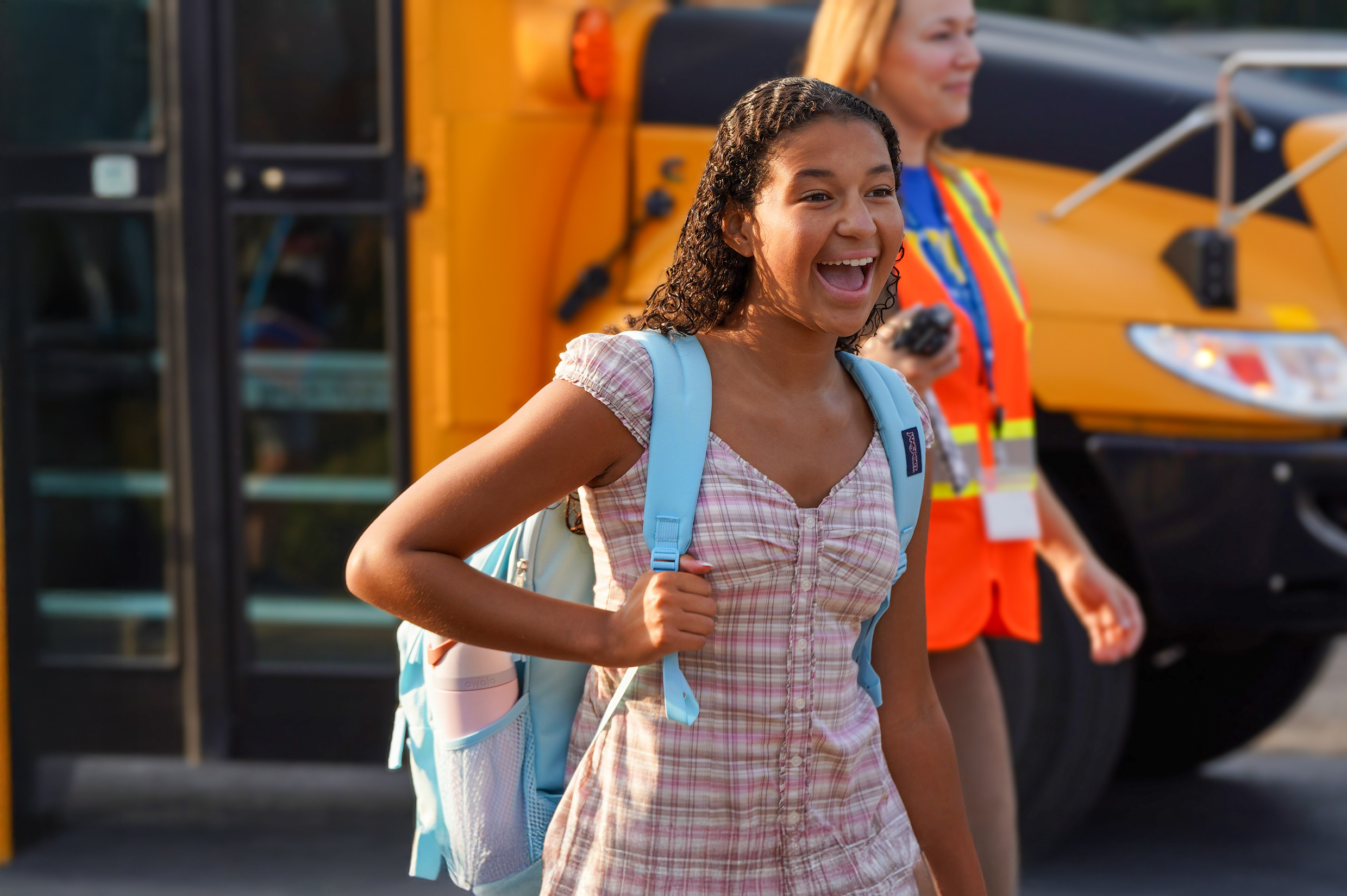 A smiling girl with a backpack walks in front of a yellow school bus where a woman in a safety vest is visible.