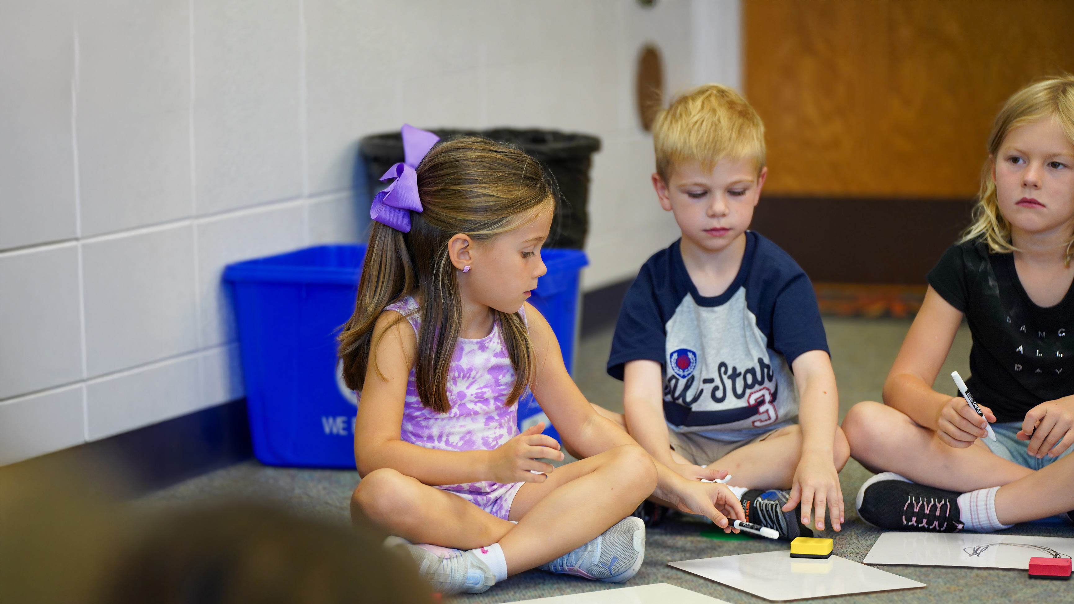 Young children sit on the floor in the foreground, engaged in an activity, while a blue recycling bin and a black trash can are visible in the background.