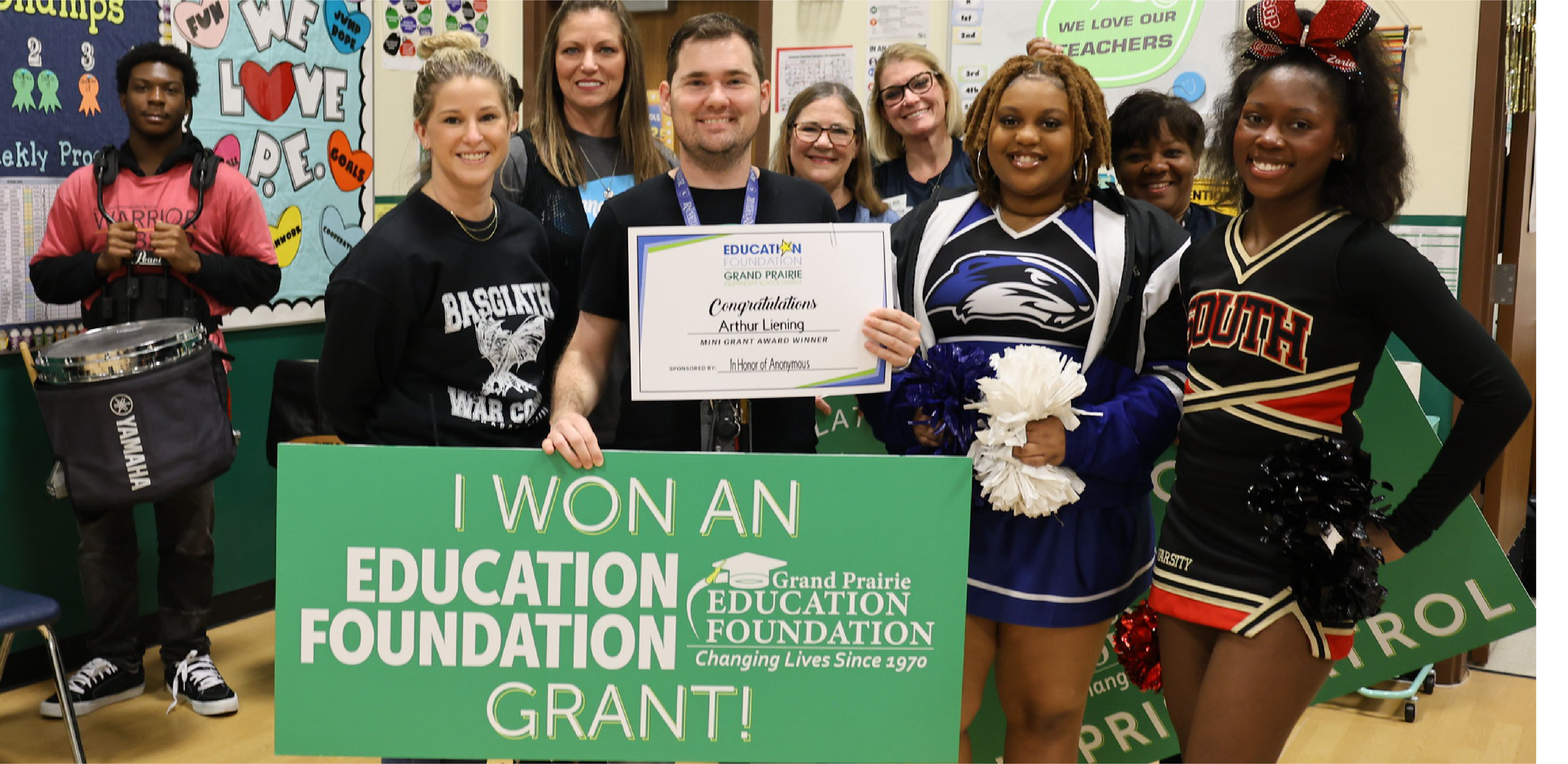 A group of smiling students and teachers stand in a classroom holding a grant award certificate and a large green sign.