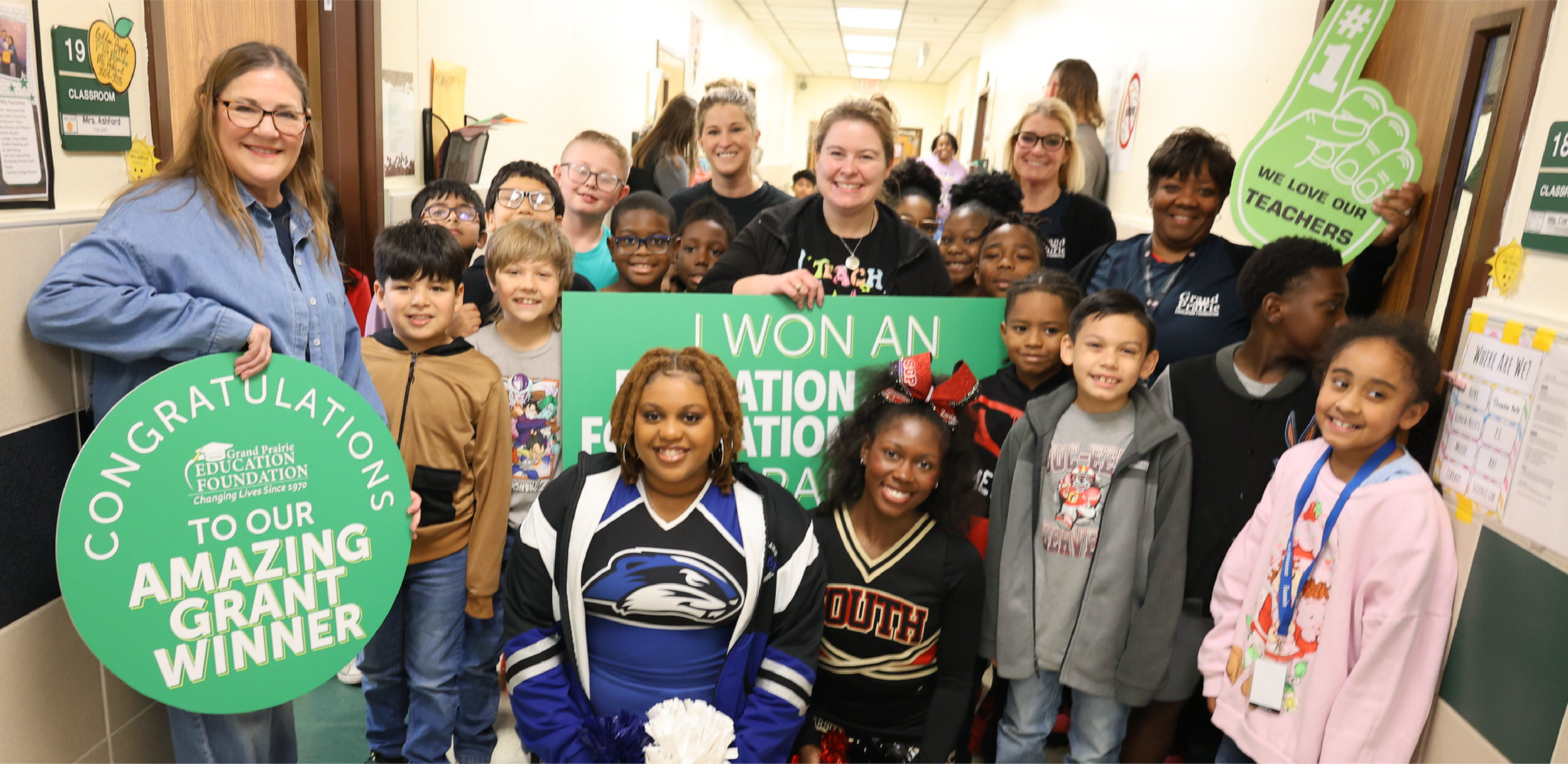 A group of smiling students and teachers pose in a school hallway, with a woman in the foreground holding a large green "Congratulations" sign.