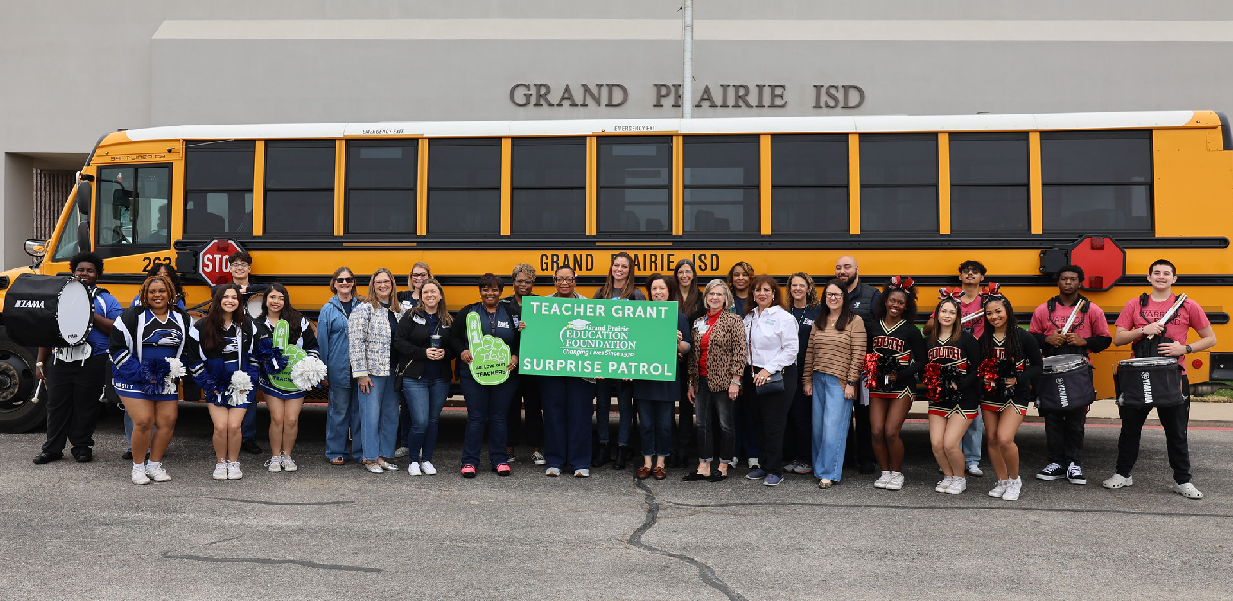 A group of people stand in the foreground holding a "Teacher Grant" sign in front of a yellow school bus and a building labeled "Grand Prairie ISD."