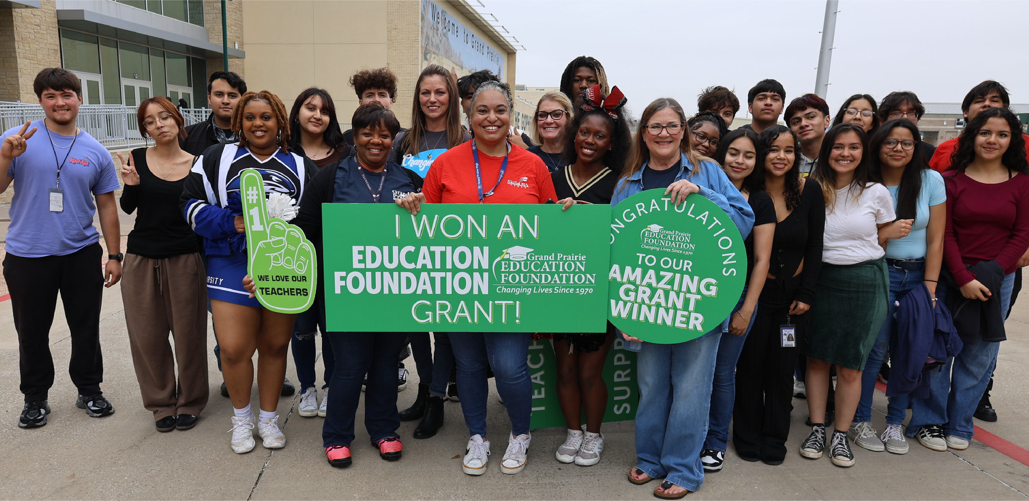 A group of smiling students and teachers hold large green celebratory signs in the foreground, standing outdoors in front of a school building.