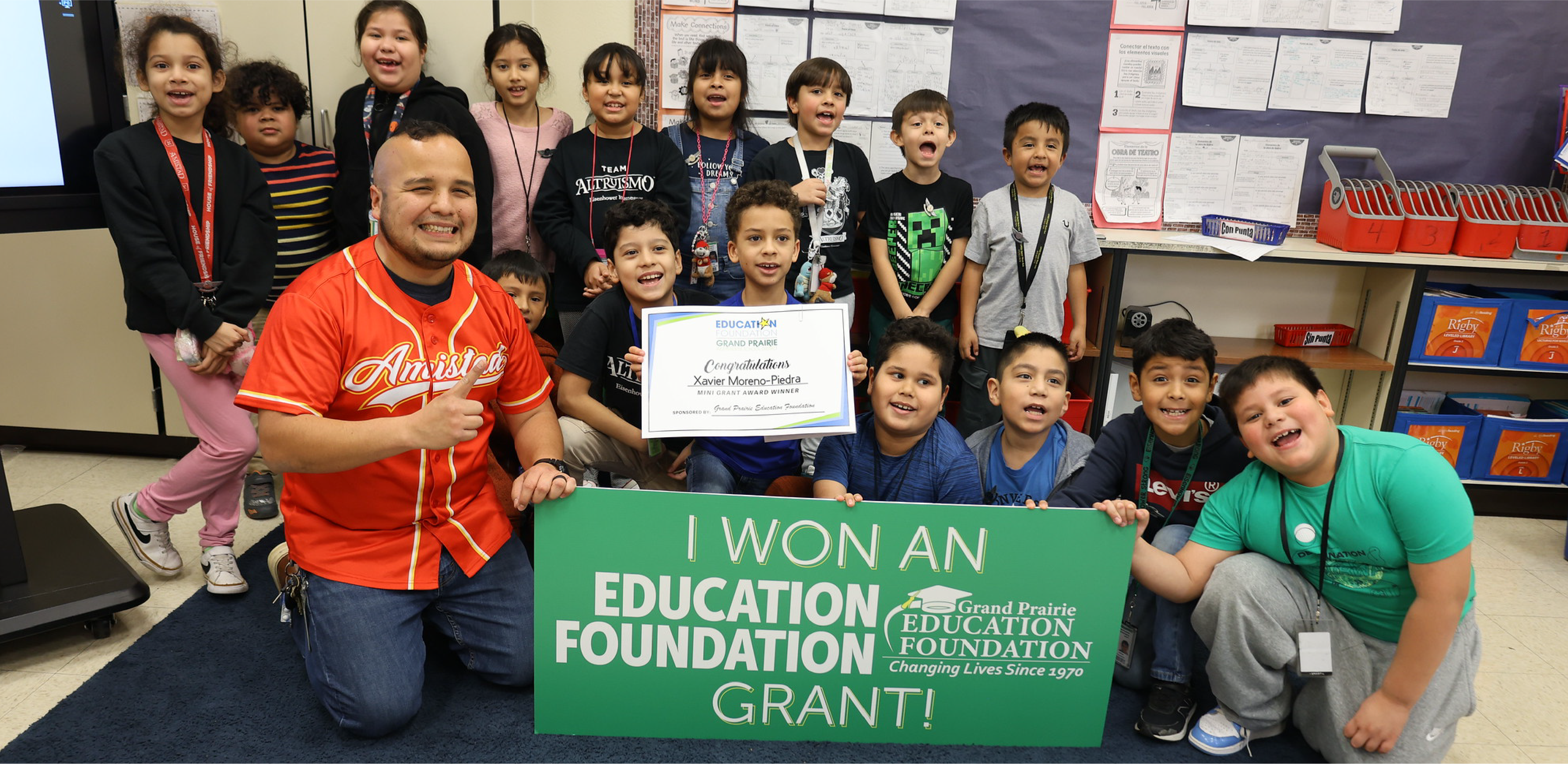 A man in a red jersey and a group of children pose with a large green grant sign in a classroom.