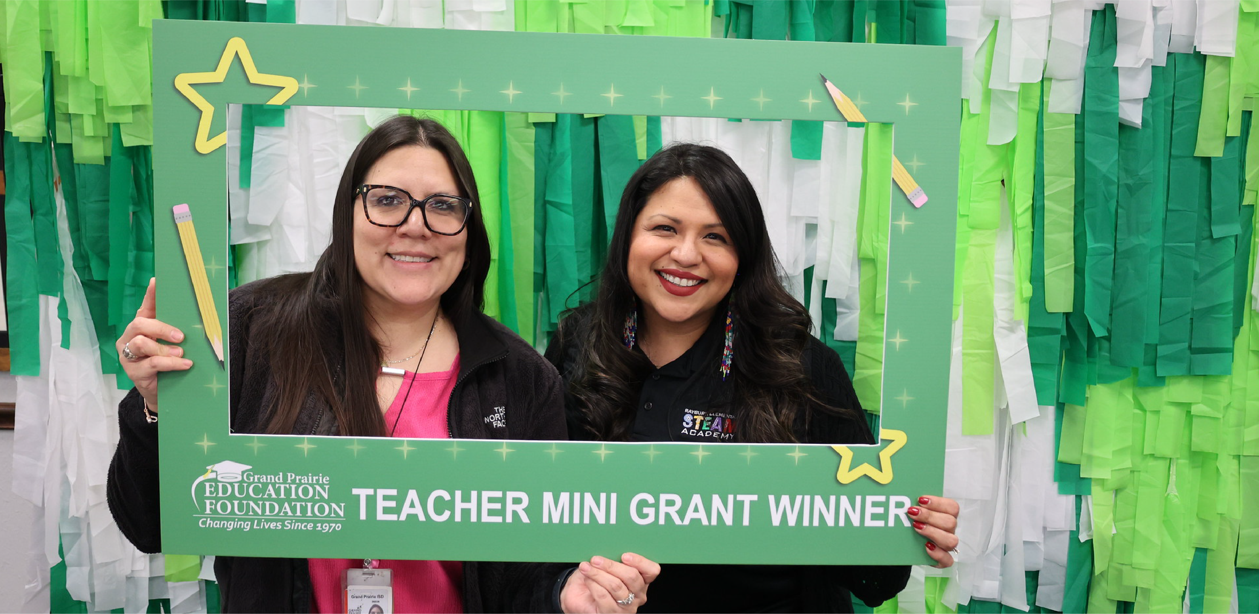 Two smiling women hold a green "Teacher Mini Grant Winner" photo frame against a background of green and white streamers.