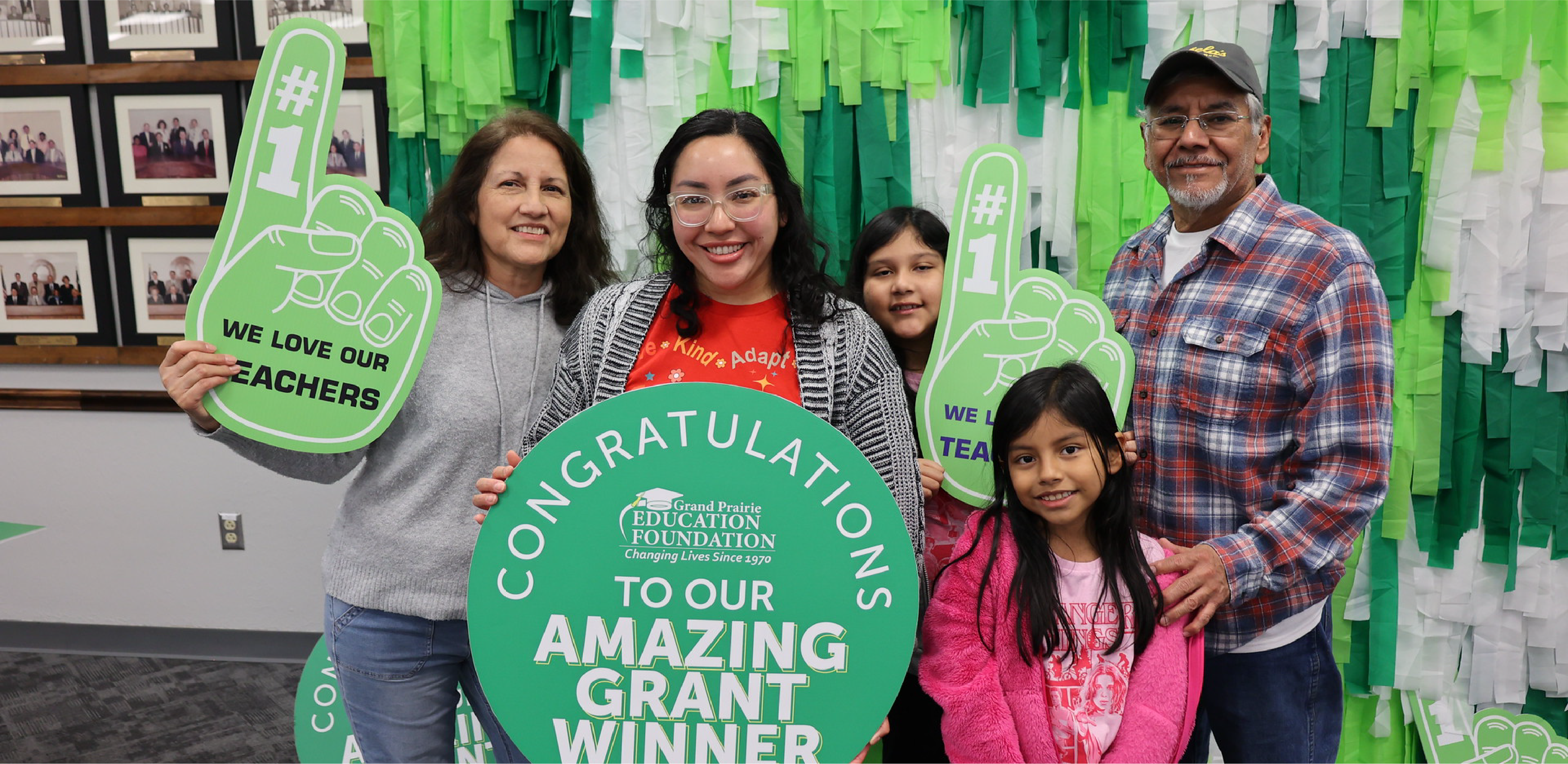 A group of people pose with celebratory signs in front of a green fringed backdrop and a wall of framed photos.