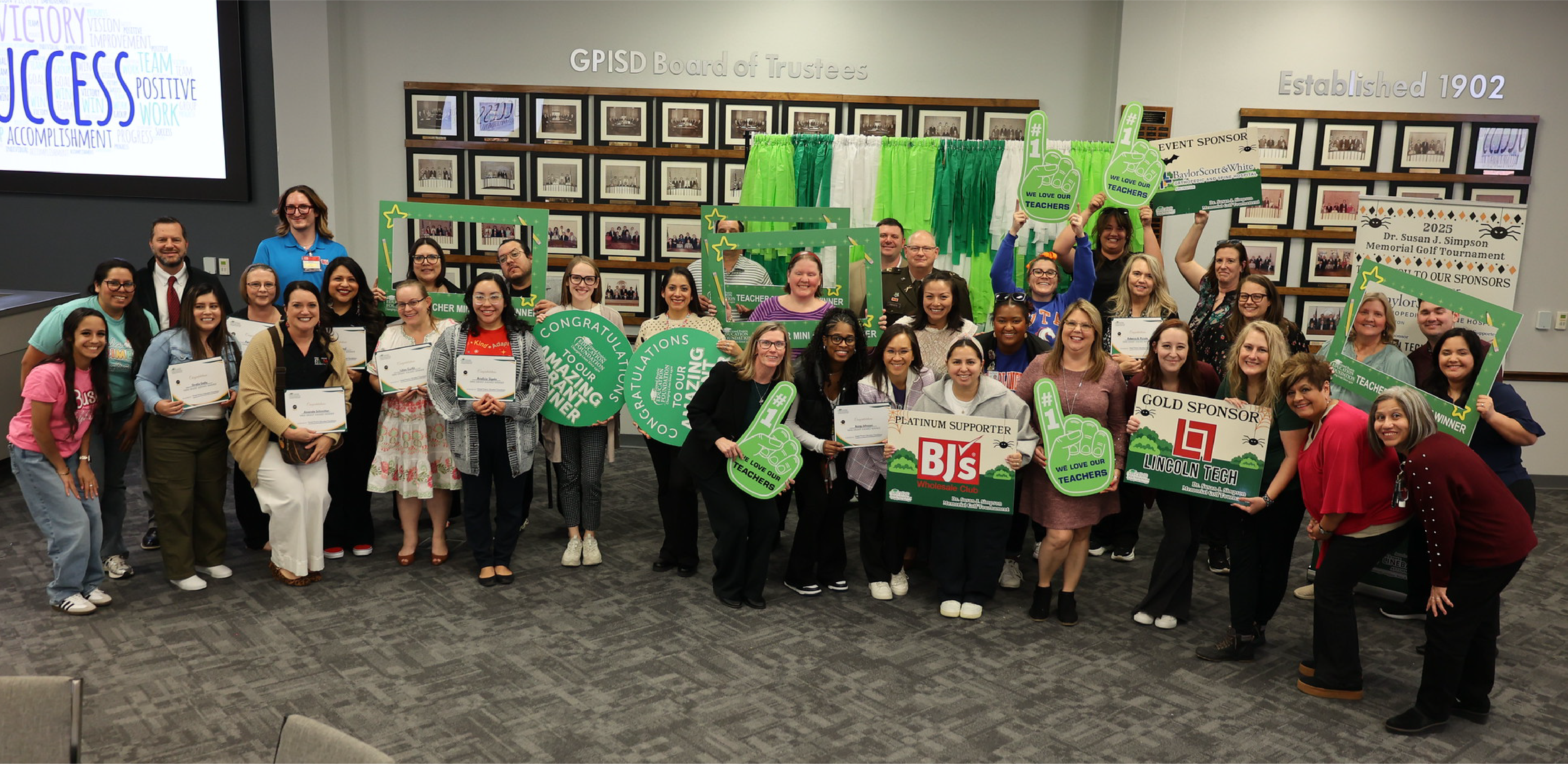 A large group of people pose together holding signs and certificates in a room with a gallery wall of framed photos.