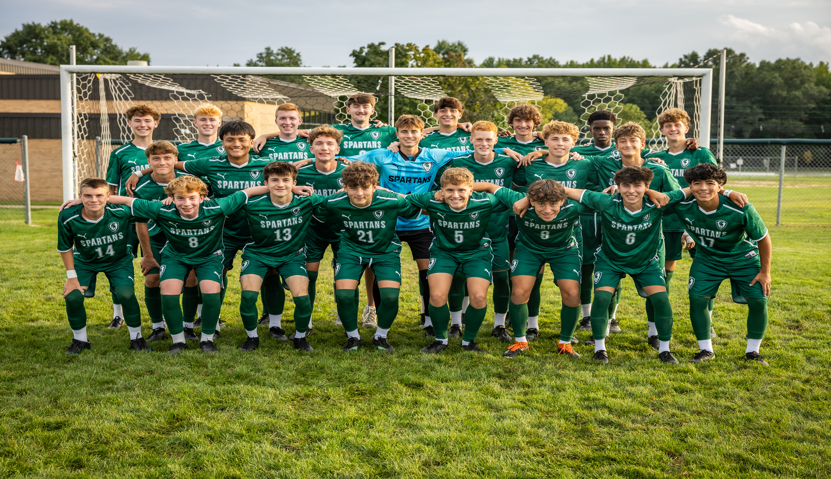 A soccer team in green uniforms poses for a photo in front of a goal on a grassy field with trees and a building in the background.