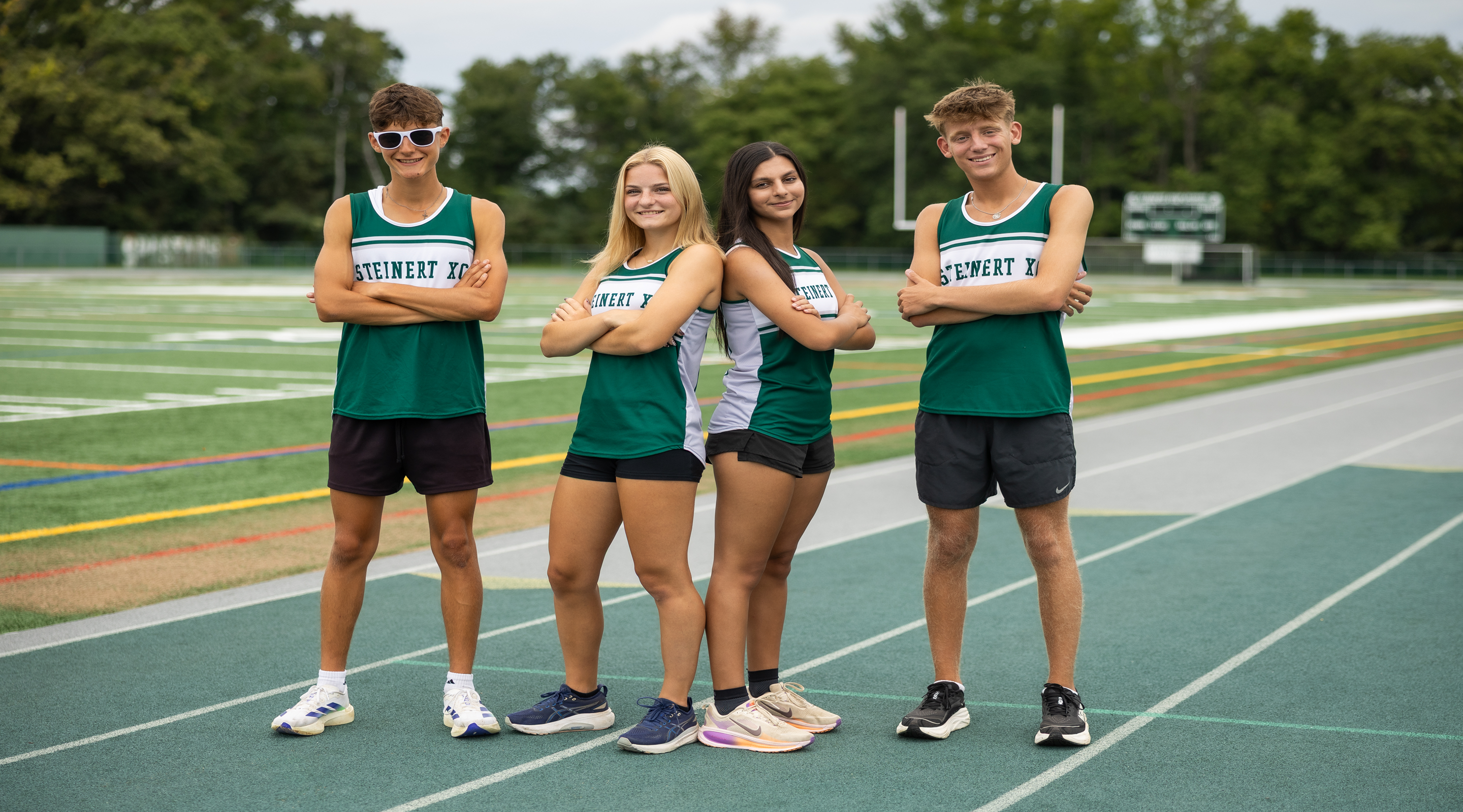 Four young athletes in green and white track uniforms stand on a track with a stadium in the background.
