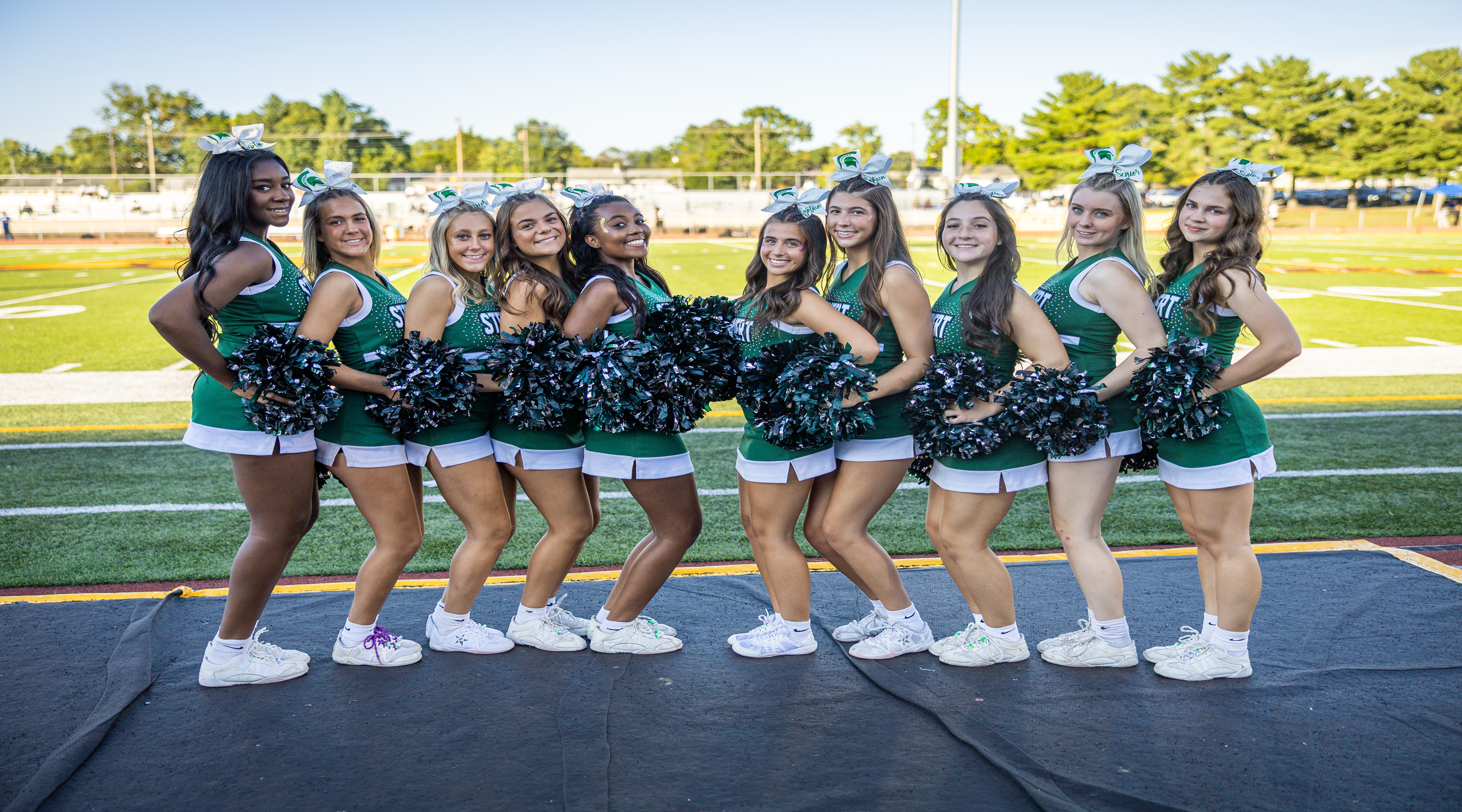 A group of nine cheerleaders in green uniforms and white sneakers stand on a dark surface in the foreground, with a football field and stadium in the background.